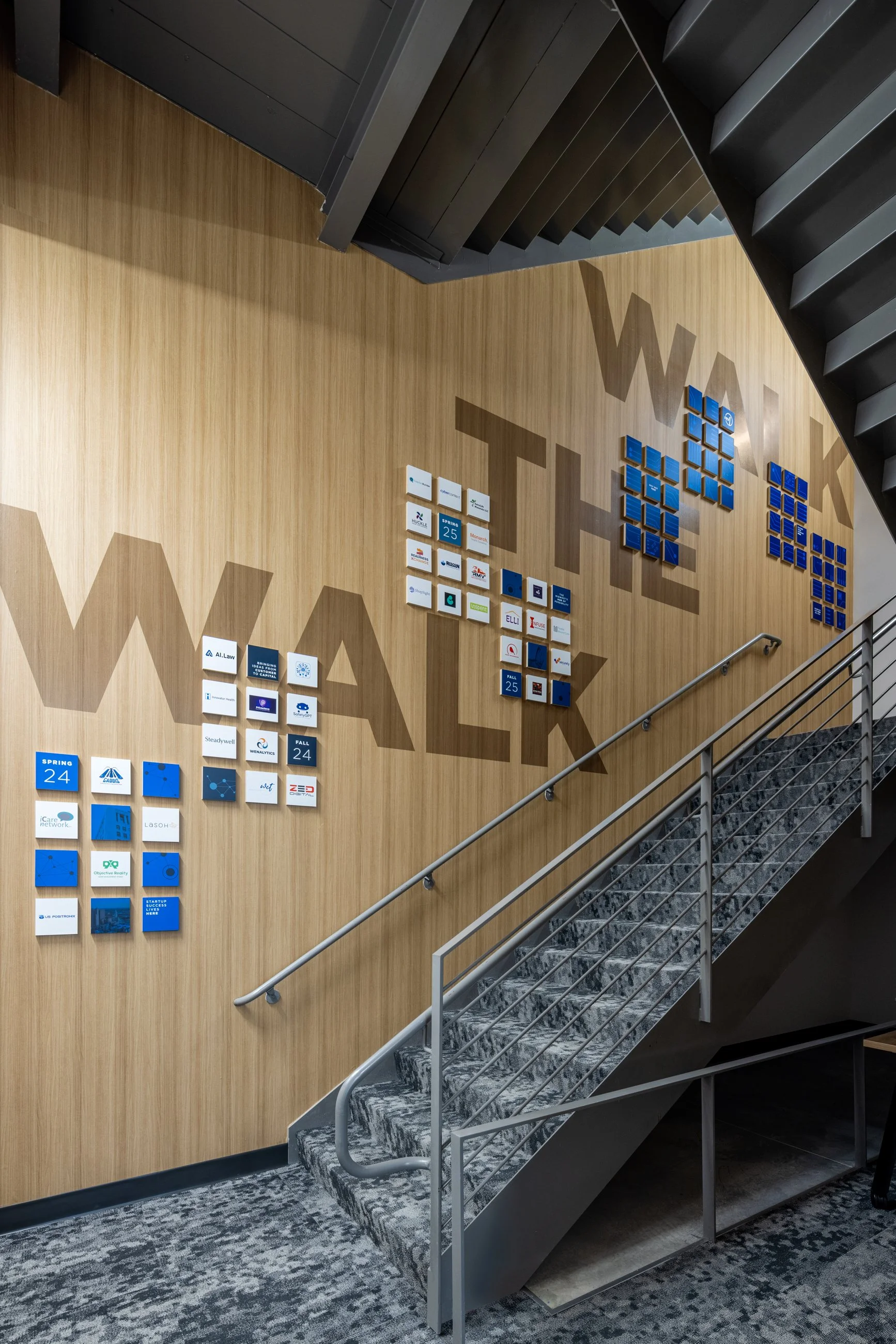 Interior stairwell with a wood-paneled wall displaying a large mural made of small square tiles, spelling out 'WALK' and 'TALK' in large letters, with a patterned carpeted floor and metal handrail.