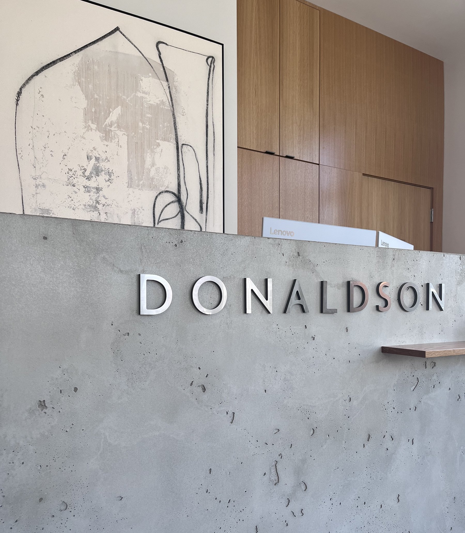 Modern reception desk with metal letters spelling 'DONALDSON' against a concrete surface, with abstract artwork on the wall and wooden cabinets in the background.