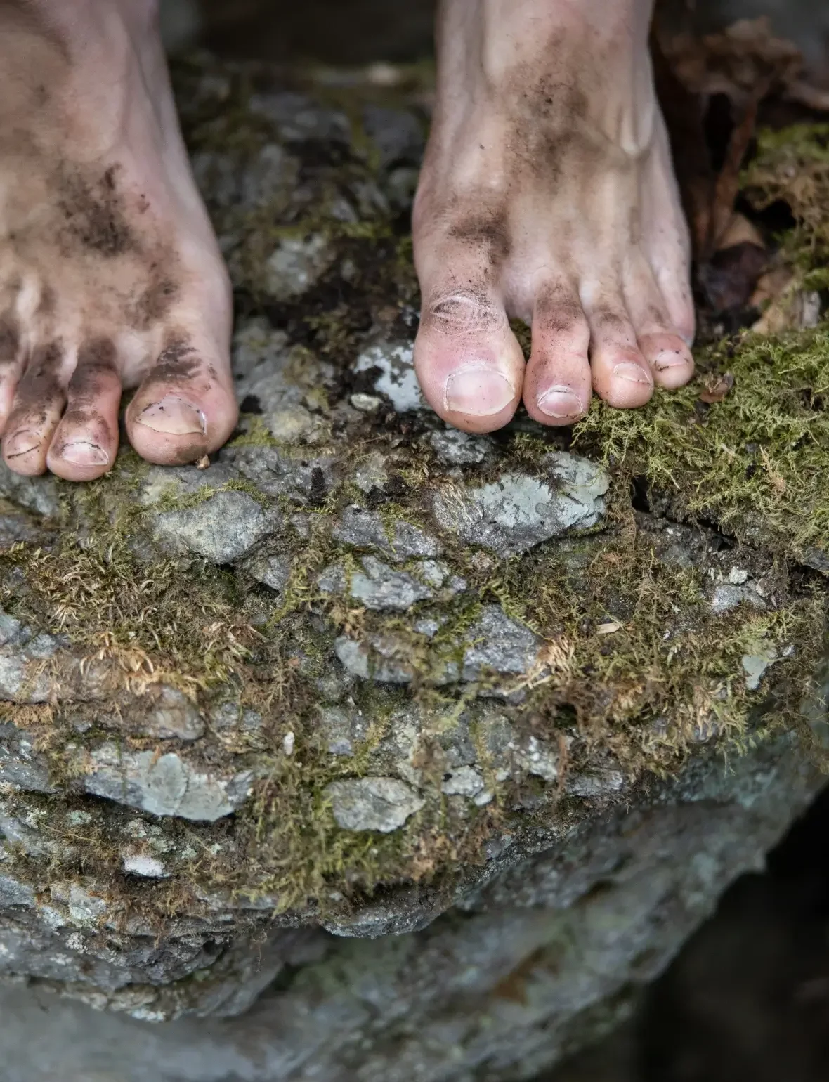 Dirty bare feet with muddy toes standing on moss and rocks outdoors.