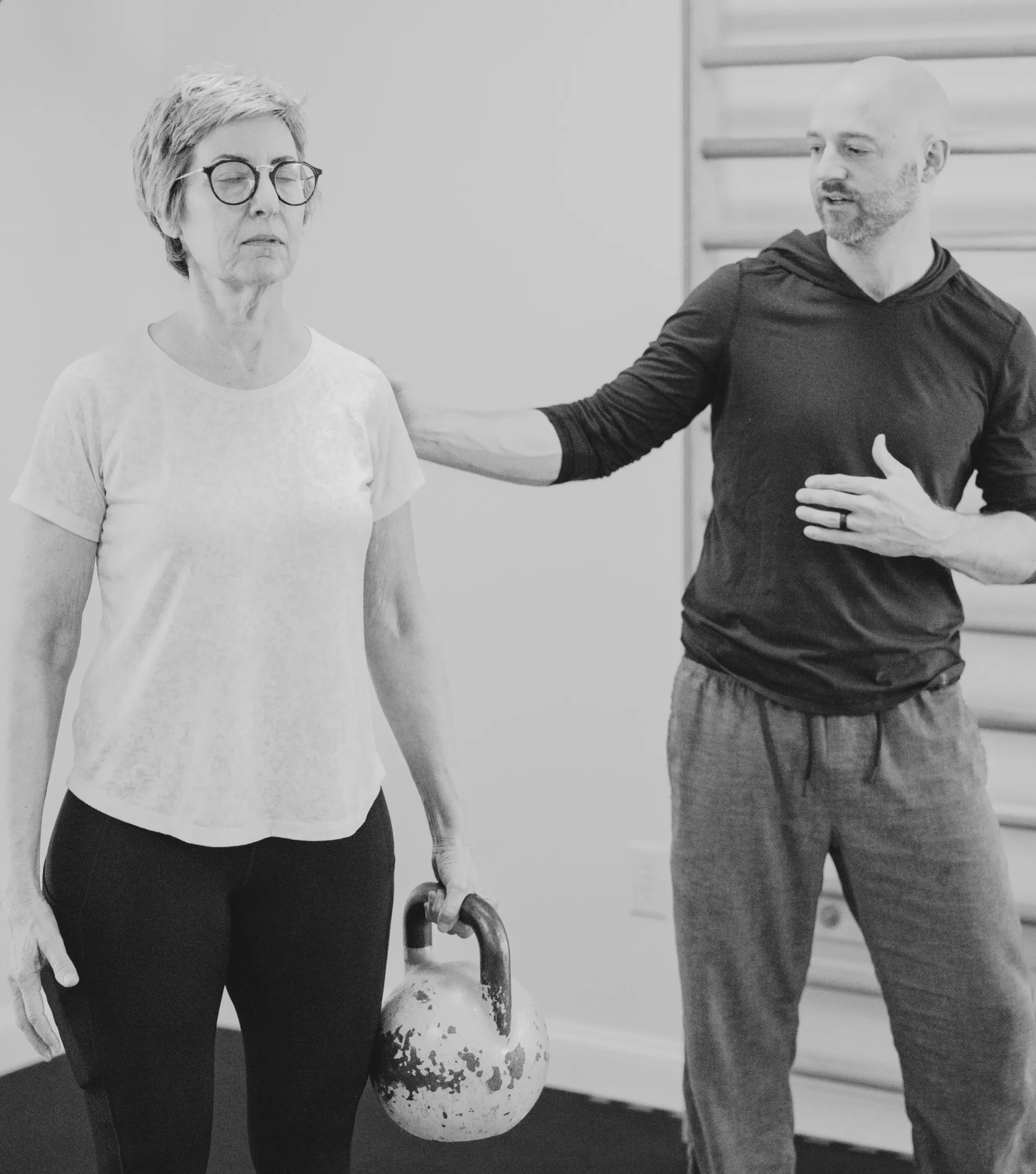 A woman with glasses and short hair holding a kettlebell in a gym, with a man beside her giving instructions.