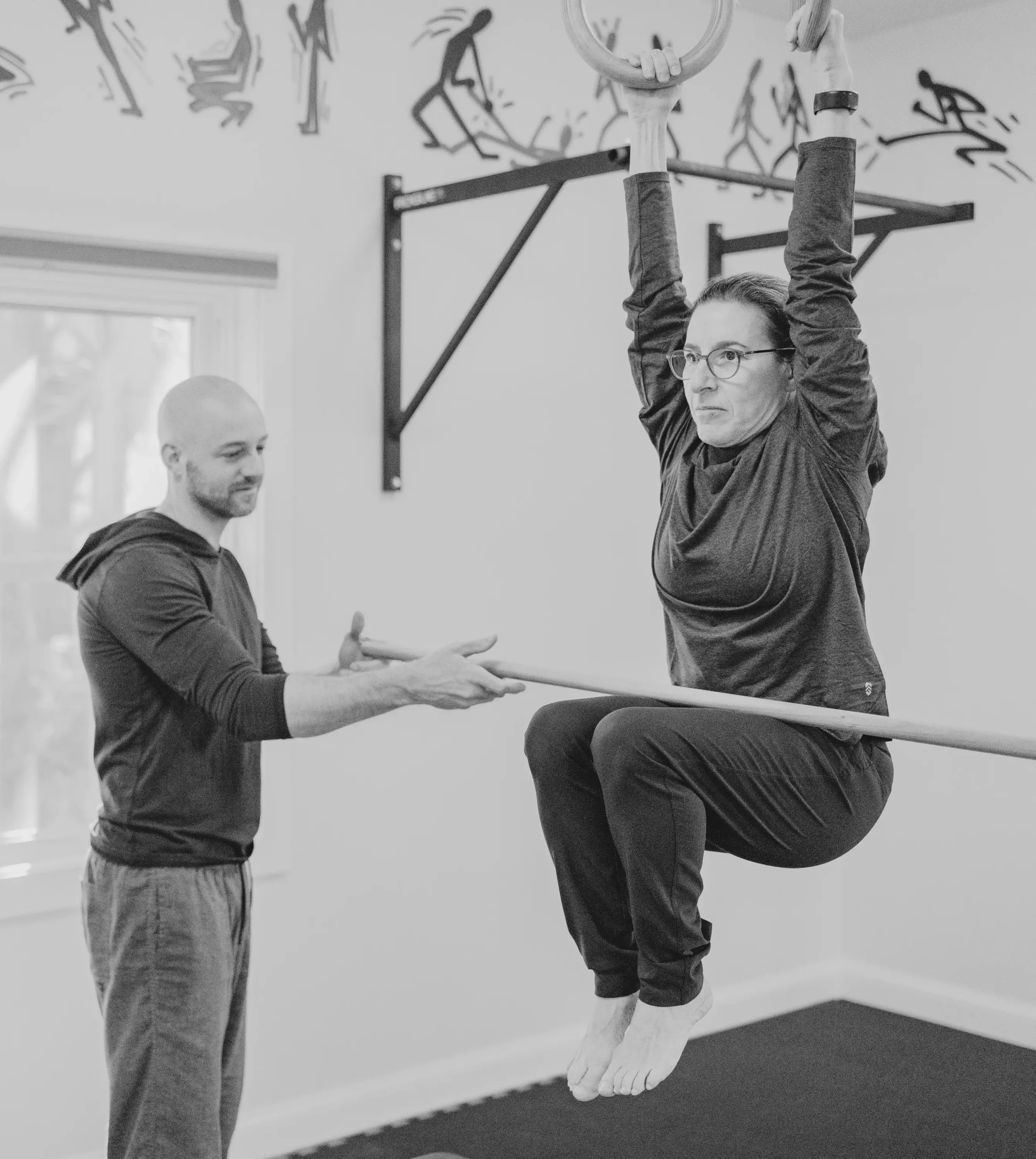 A woman with glasses and a dark long-sleeve shirt is performing a pull-up exercise on gym rings, with her knees bent and feet off the ground. A man with a bald head and beard, wearing a long-sleeve shirt, is assisting her by holding a bar underneath her.