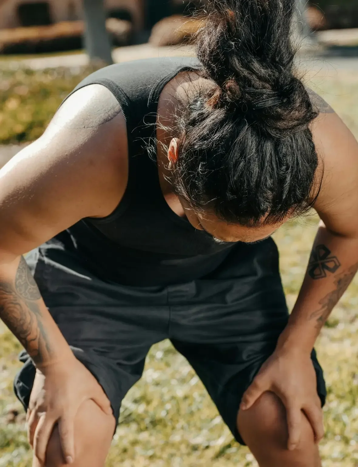 A woman with tattoos on her arms wearing a black tank top and shorts, hunched over outdoors in sunlight, possibly resting or catching her breath.