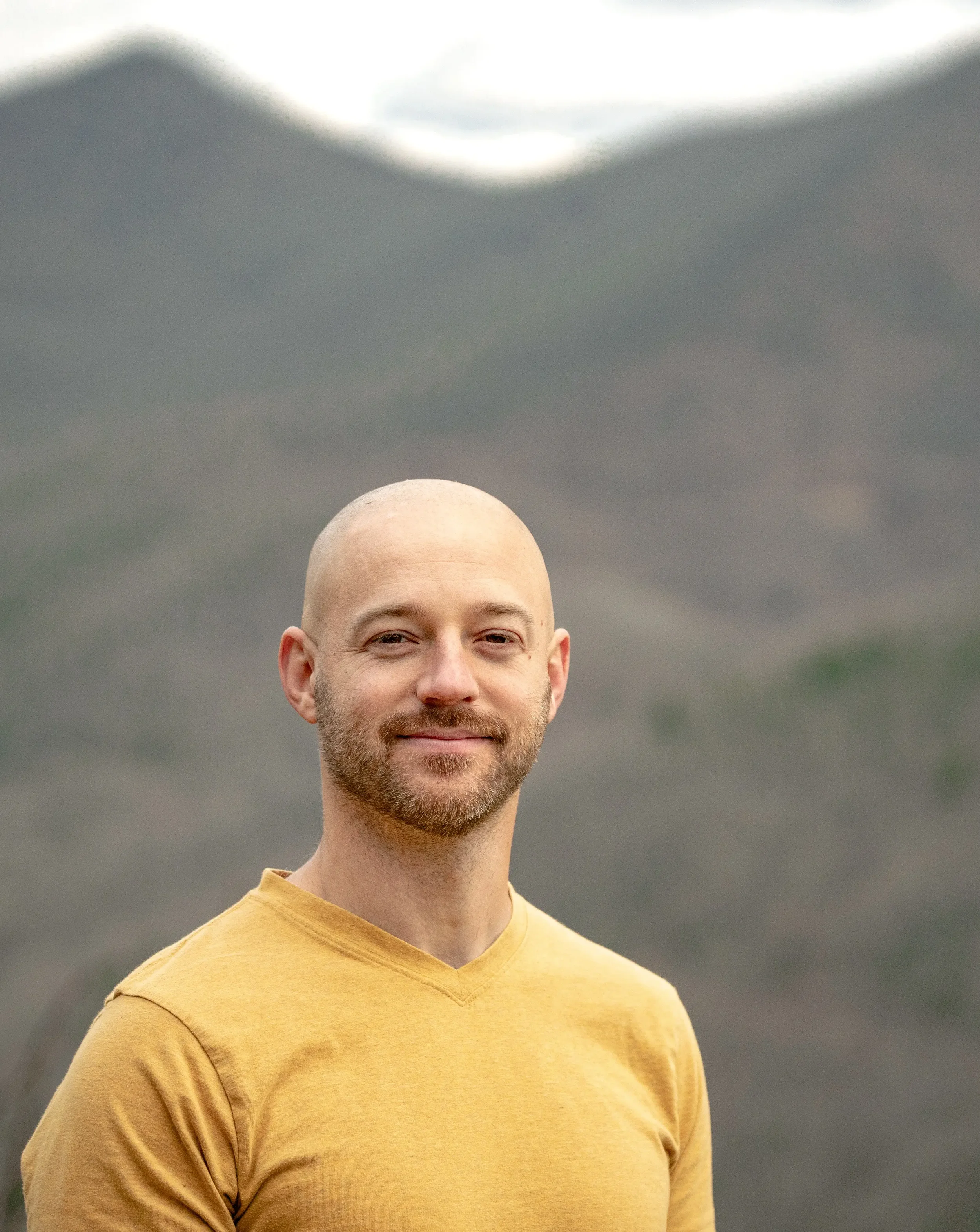 A man with a beard and buzz cut, wearing a mustard yellow shirt, standing outdoors with a blurred mountainous landscape in the background.