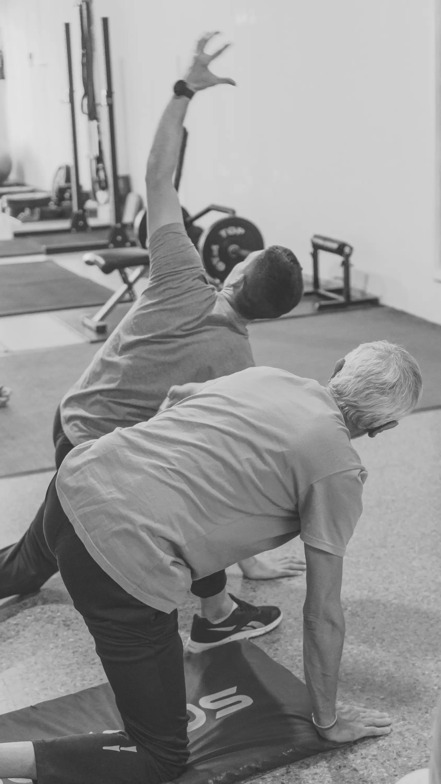 Two men performing stretching exercises in a gym, one stretching on the ground with hands on the floor and the other with one arm raised overhead.