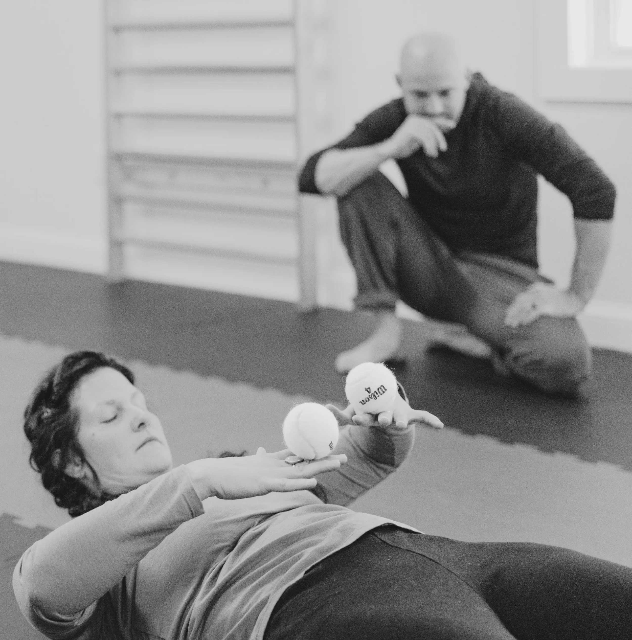 A woman lying on the floor holding two tennis balls, with a man kneeling nearby in a gym or therapy room.