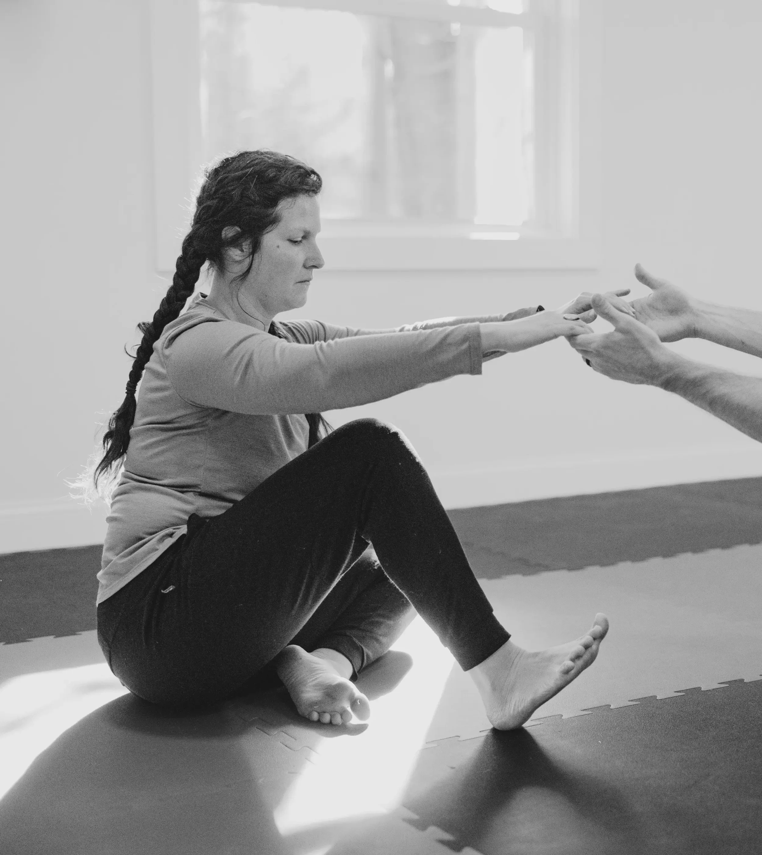 A woman with long dark hair braided, sitting cross-legged on a mat, stretching her arms forward while a person in the background helps her extend her reach.