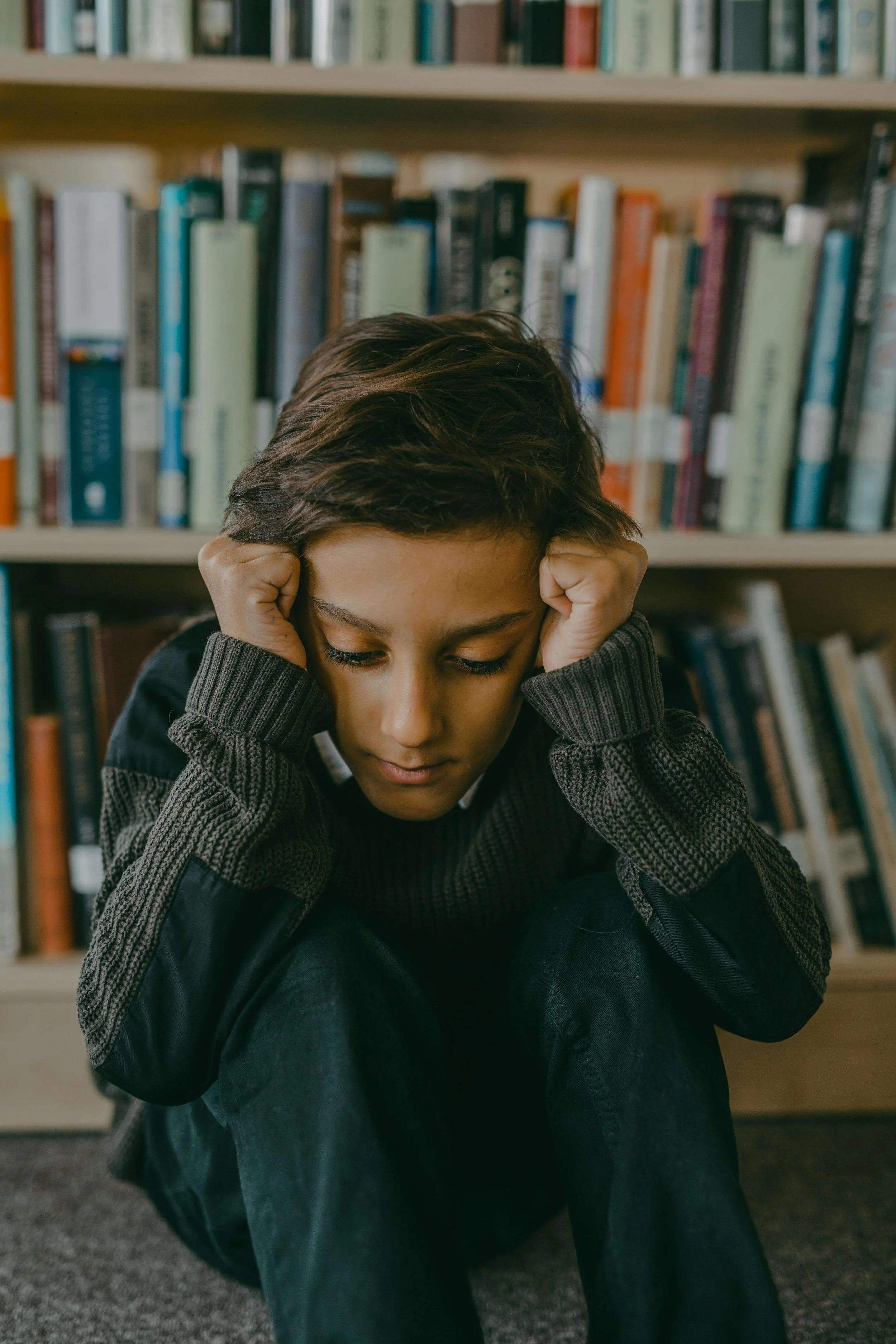 Child sitting alone with head down in front of bookshelves. Child counseling in Columbia, MO supports children who feel overwhelmed by anxiety, helping them develop coping skills and emotional resilience.