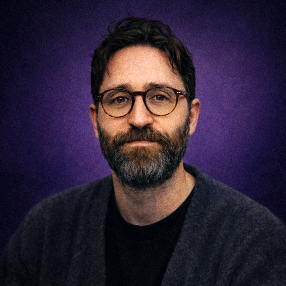 A man with glasses, a beard, and dark hair, wearing a dark sweater, looking at the camera against a purple background.