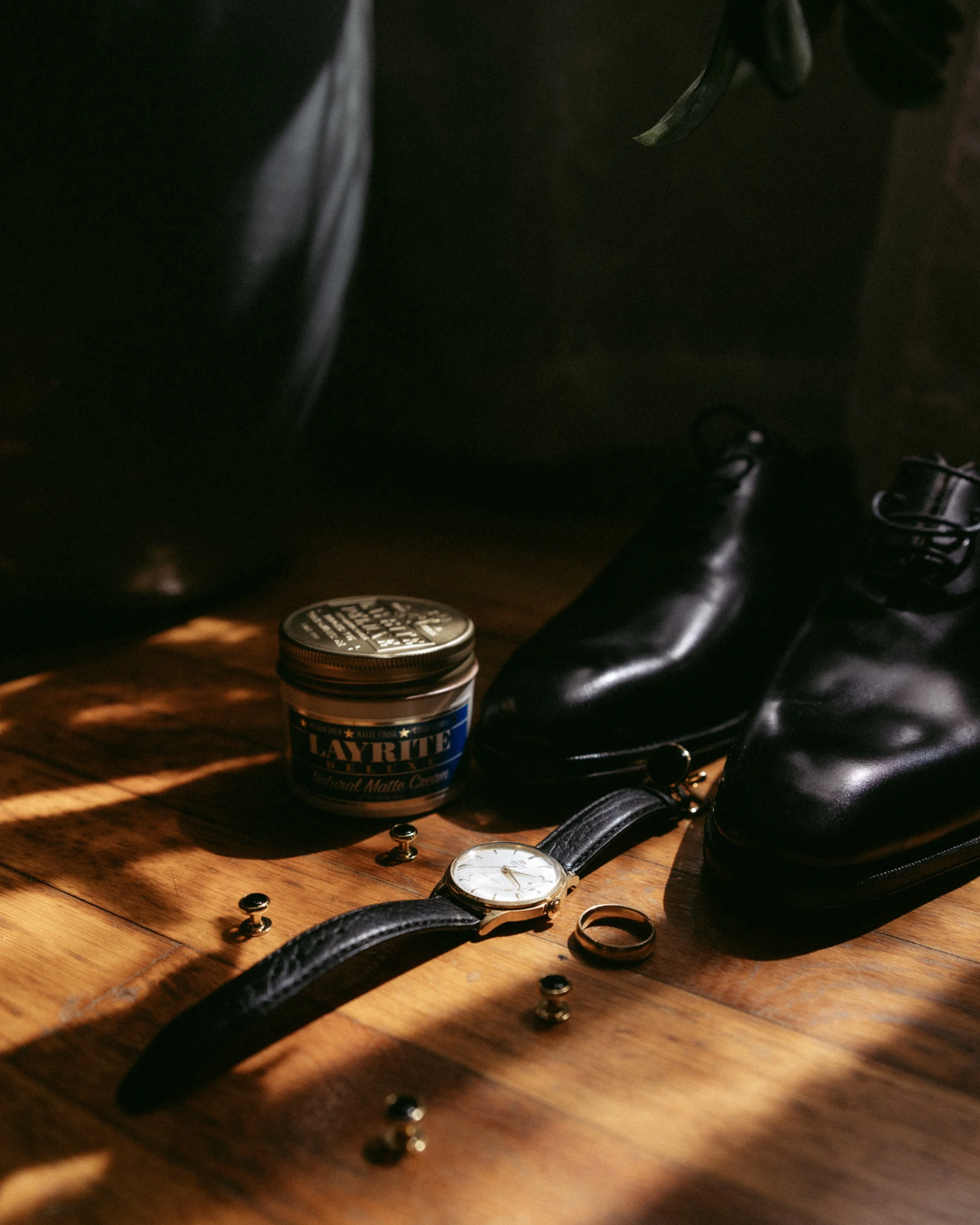 The image shows a wooden surface with various objects including a leather belt, wristwatch, a small jar of Layrite matte cream, a pair of black shoes, and scattered metal studs.