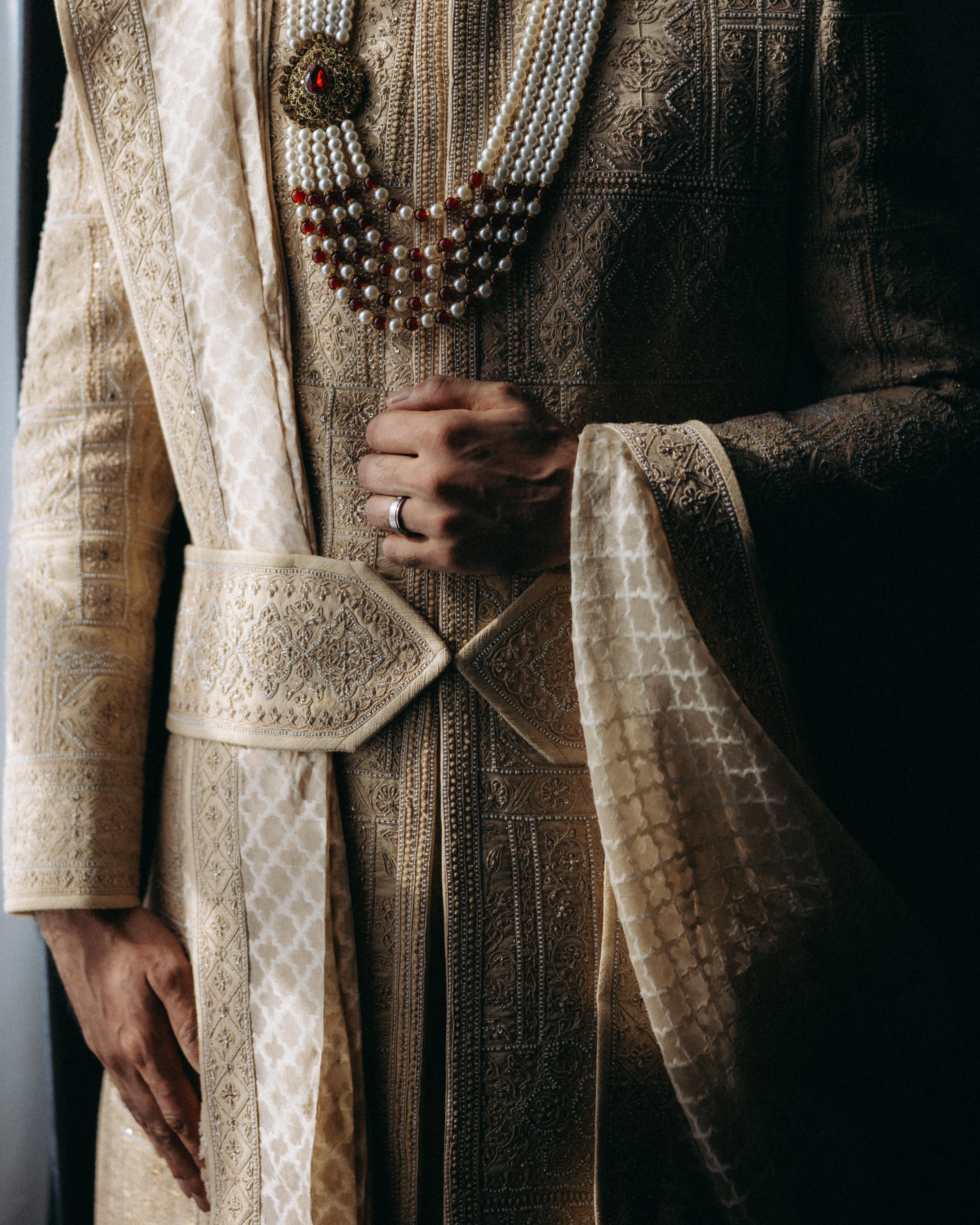 Close-up of a person wearing traditional Indian attire with detailed embroidery and jewelry, including multiple layers of pearl and red gemstone necklaces, and a gold and silver ornate fabric.