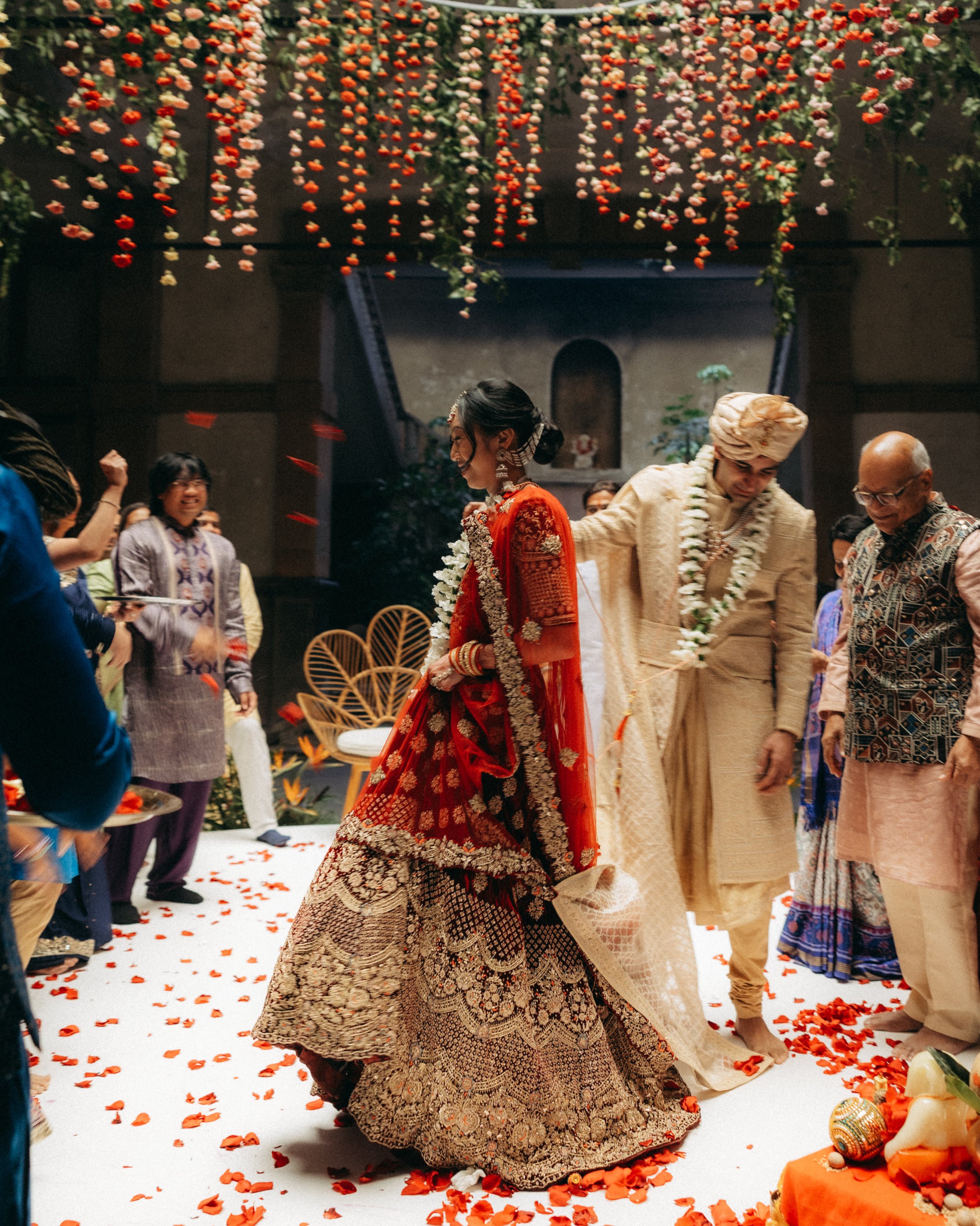 Indonesian wedding ceremony with bride and groom in traditional attire, surrounded by guests, floral decorations, and a white floral carpet.