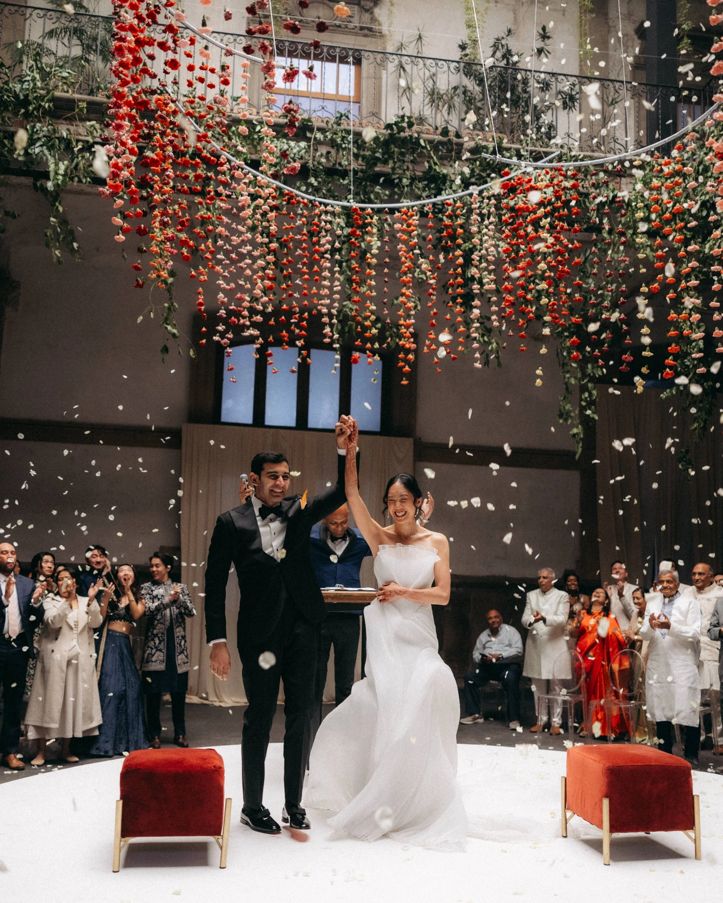 A wedding celebration where a bride and groom are standing together, smiling, and surrounded by guests. The bride is wearing a white wedding dress and the groom is in a black tuxedo. There are colorful flower arrangements hanging above and white flow