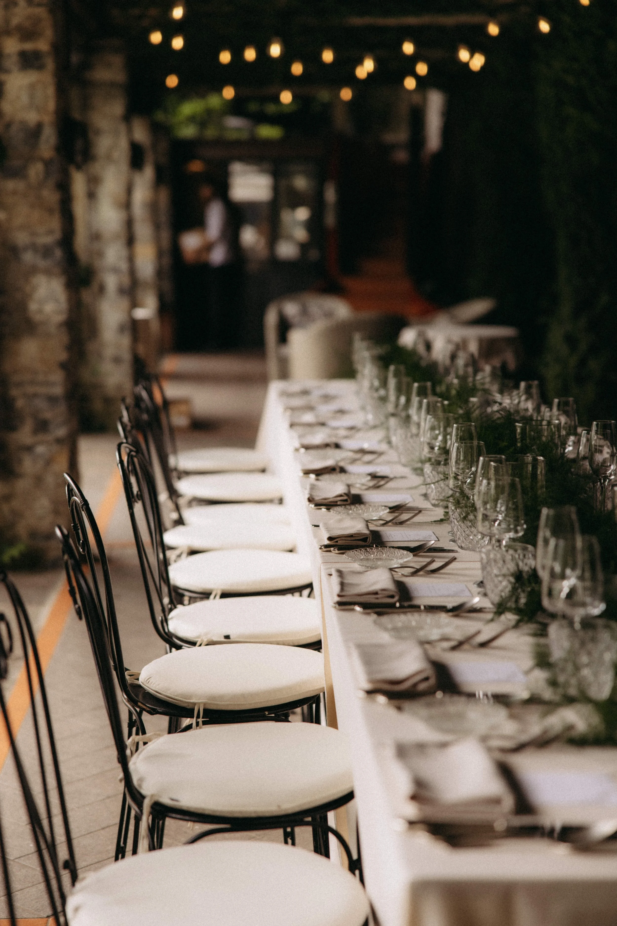 A long banquet table set for a formal event, with white tablecloths, glassware, silverware, and napkins, in a cozy, dimly lit space with string lights and rustic brick walls.