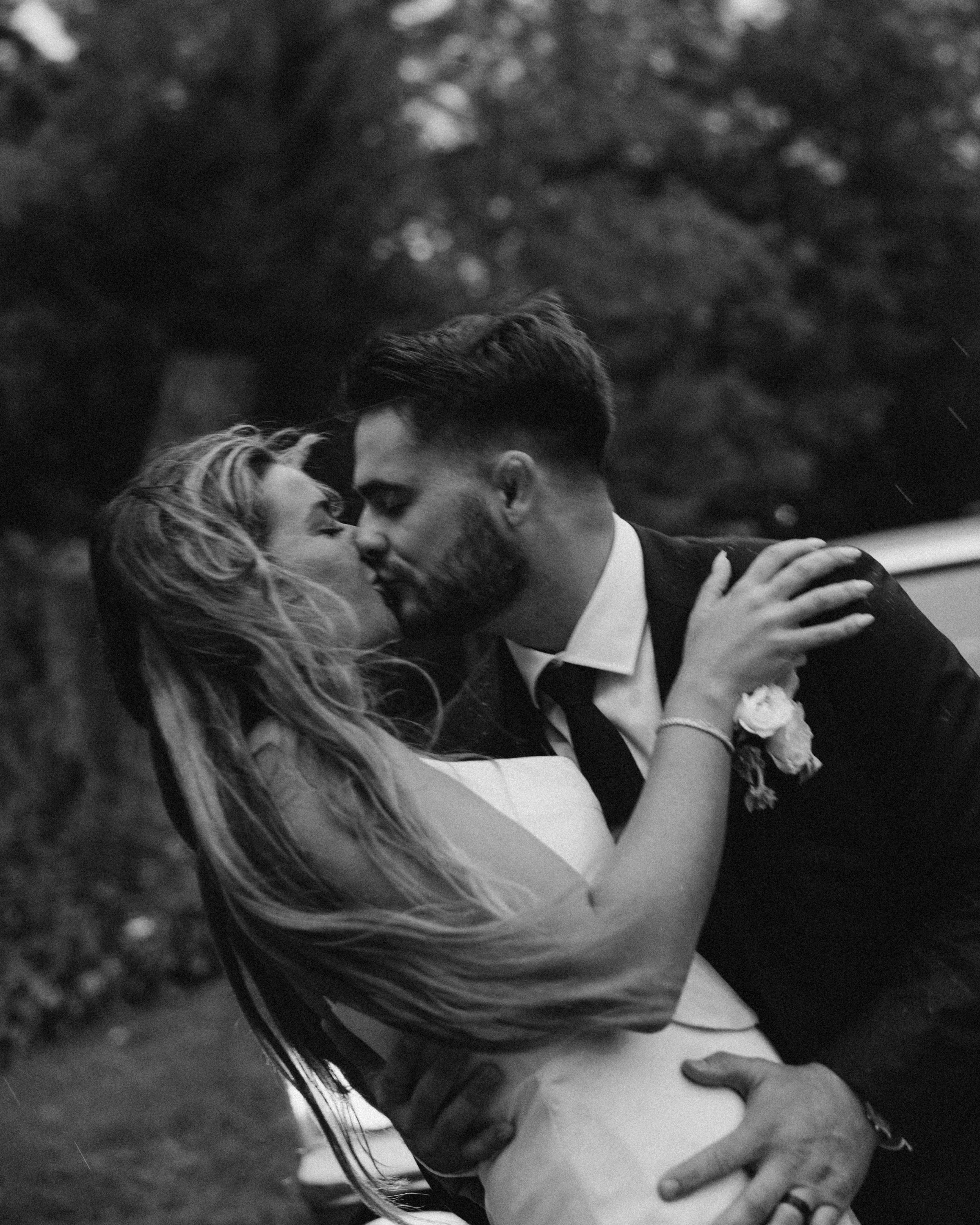 A black and white photo of a couple kissing outdoors. The woman is wearing a sleeveless dress, and the man is dressed in a suit. They are embracing each other closely with their eyes closed.