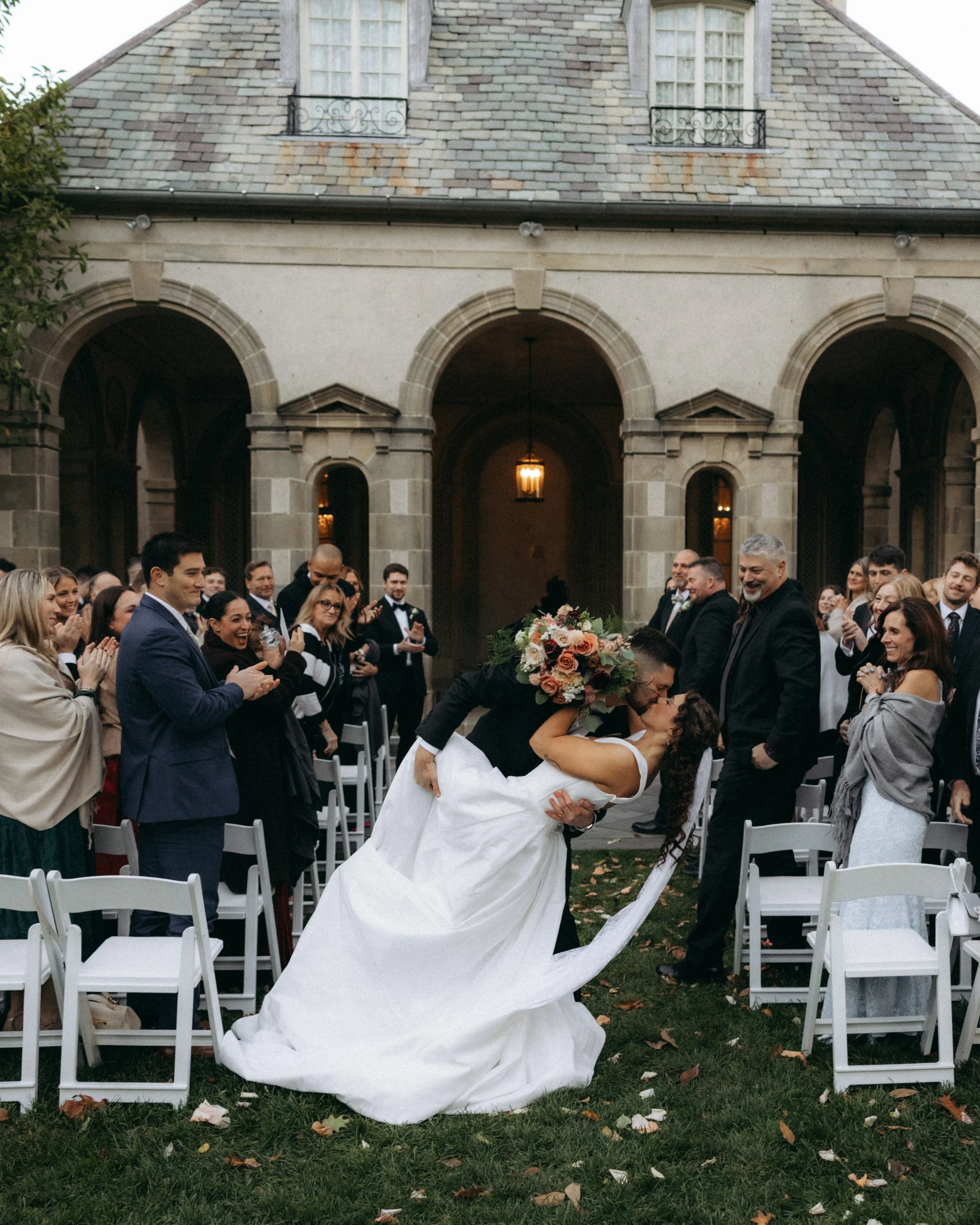 A bride and groom sharing a kiss during their wedding ceremony outdoors, with guests seated in white chairs and a stone building with arched doorways and windows in the background.