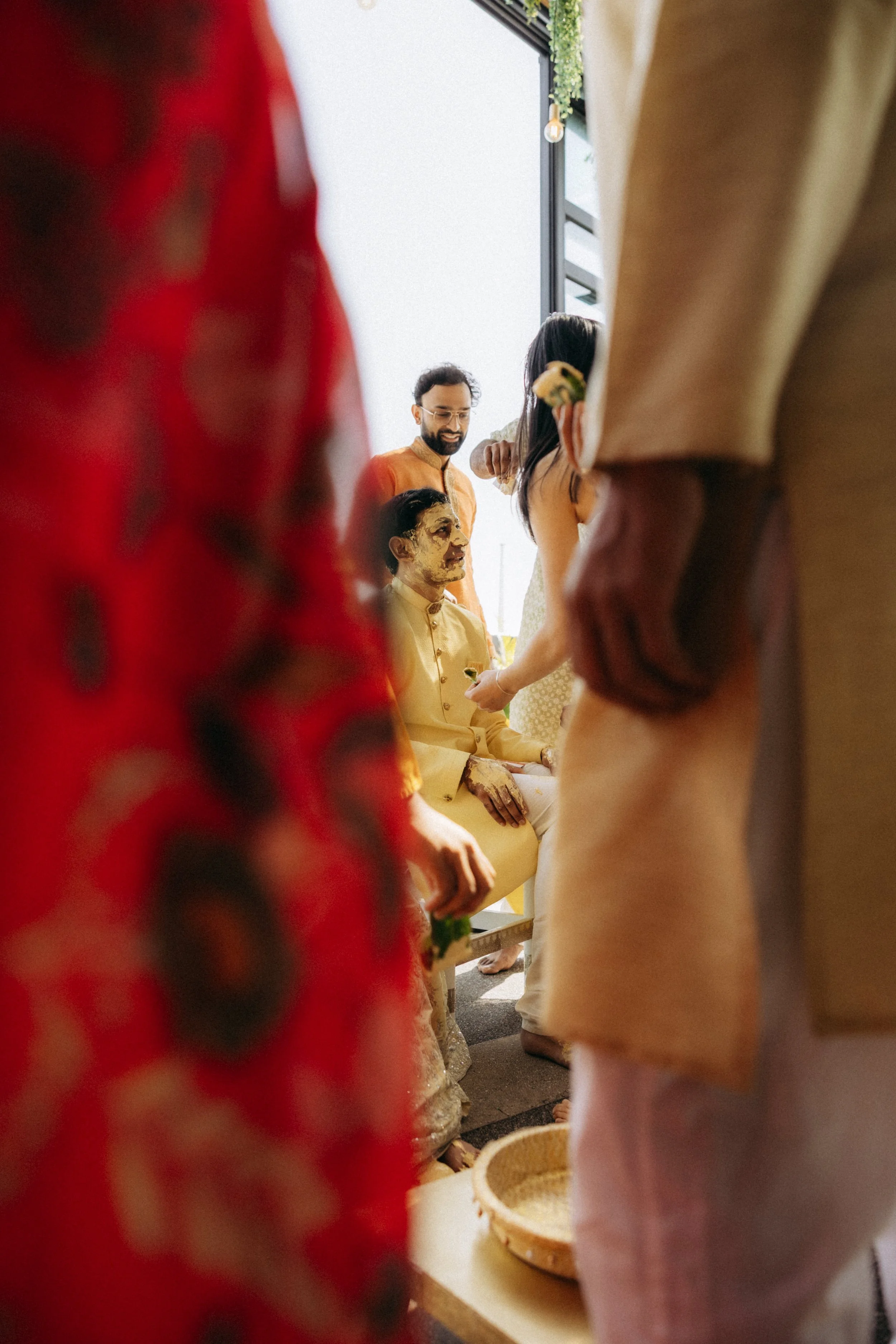 People participating in a traditional Indian pre-wedding ceremony, with men wearing kurta and women in sarees, inside a decorated space with sunlight coming through the window.