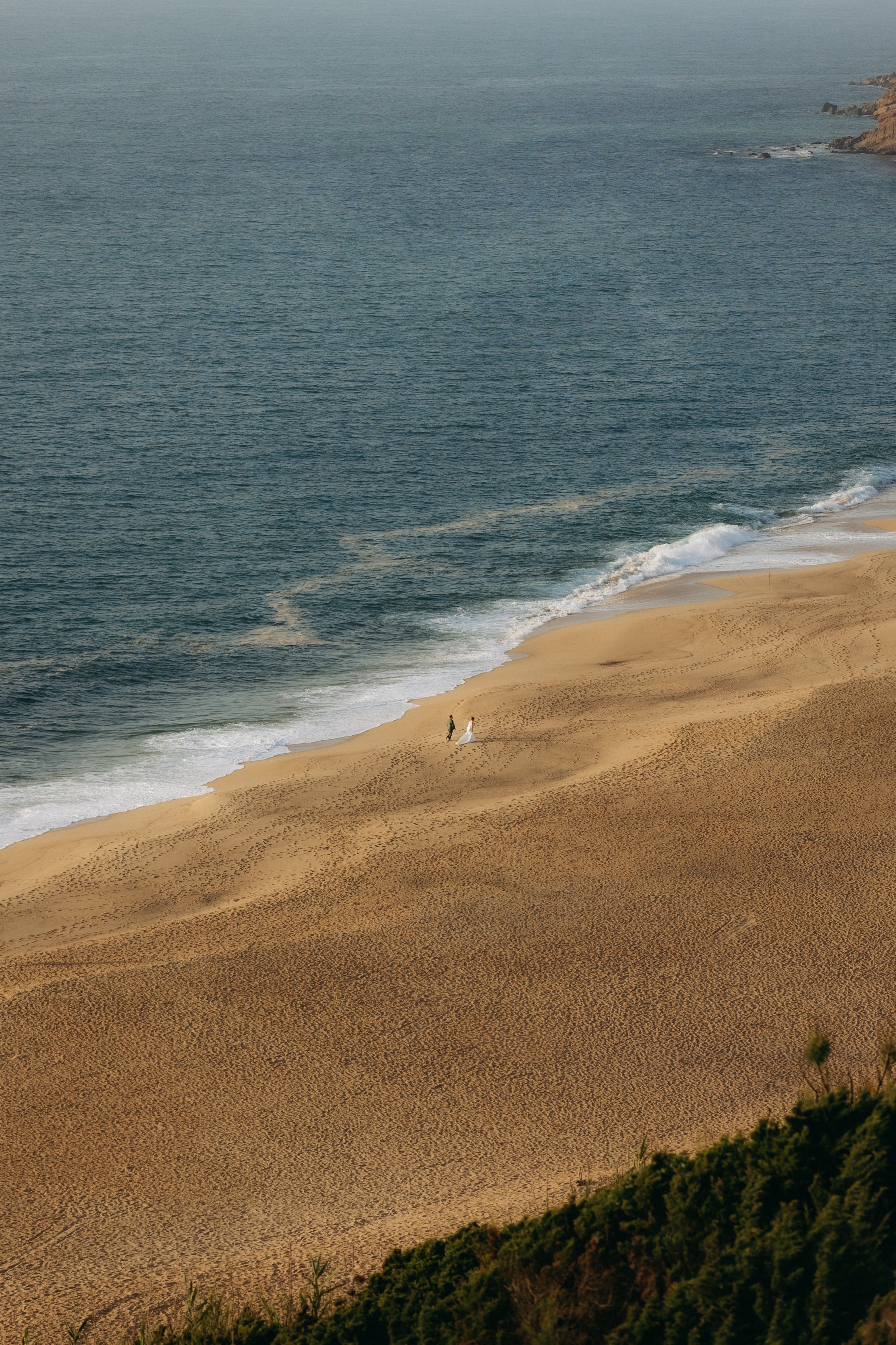 A sandy beach with gentle waves and a few people walking near the shoreline, bordered by green bushes and rocky cliffs in the distance.