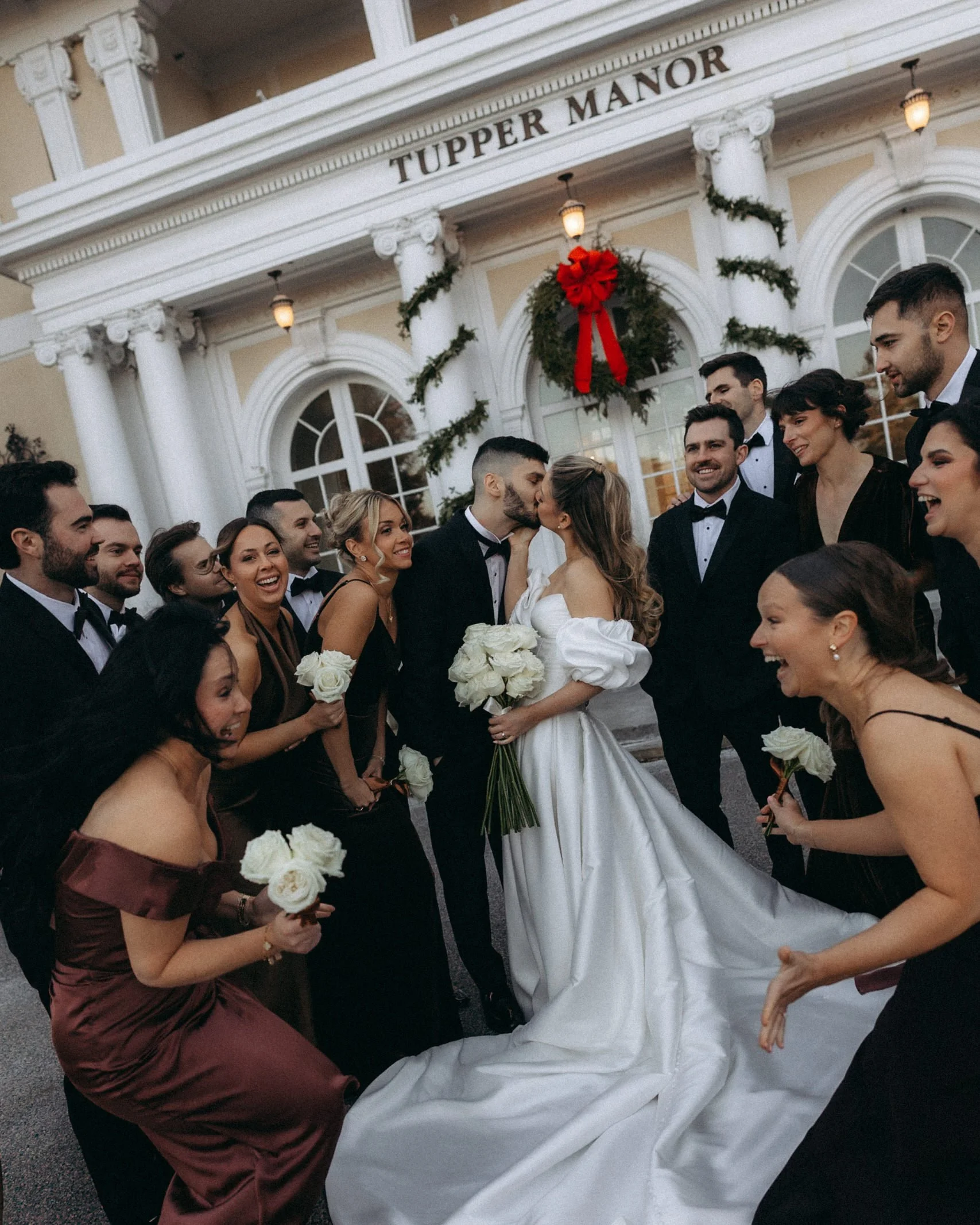 Bride and groom kissing surrounded by wedding guests outside of a decorated building with 