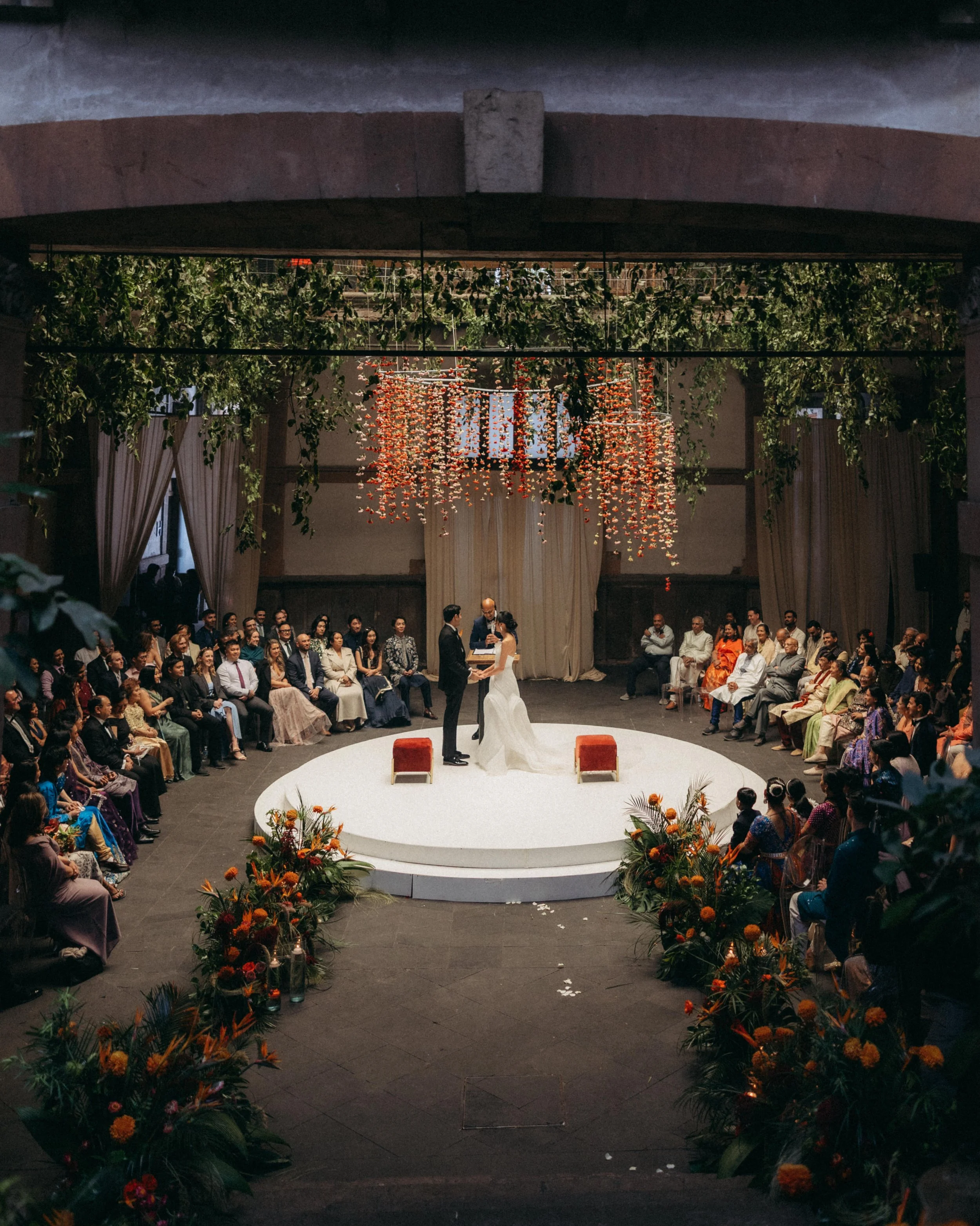 A wedding ceremony taking place indoors, with a couple standing in front of a officiant on a white circular platform surrounded by floral arrangements. Guests are seated around the platform, and decorations include hanging floral installations from t