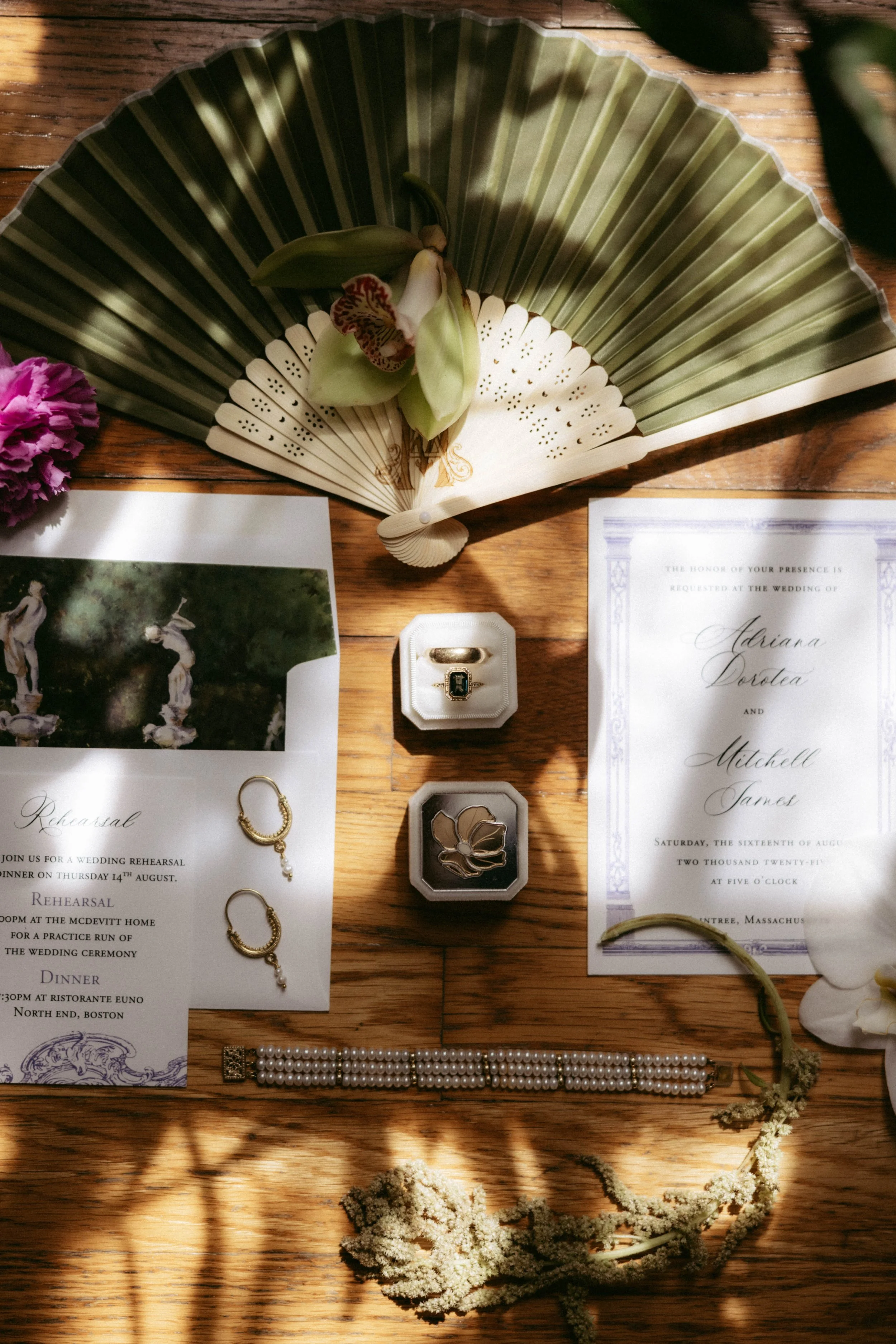 Wedding invitation suite displayed on a wooden table with a green fan and various jewelry pieces, including pearl necklaces, rings, and earrings, along with floral accents.