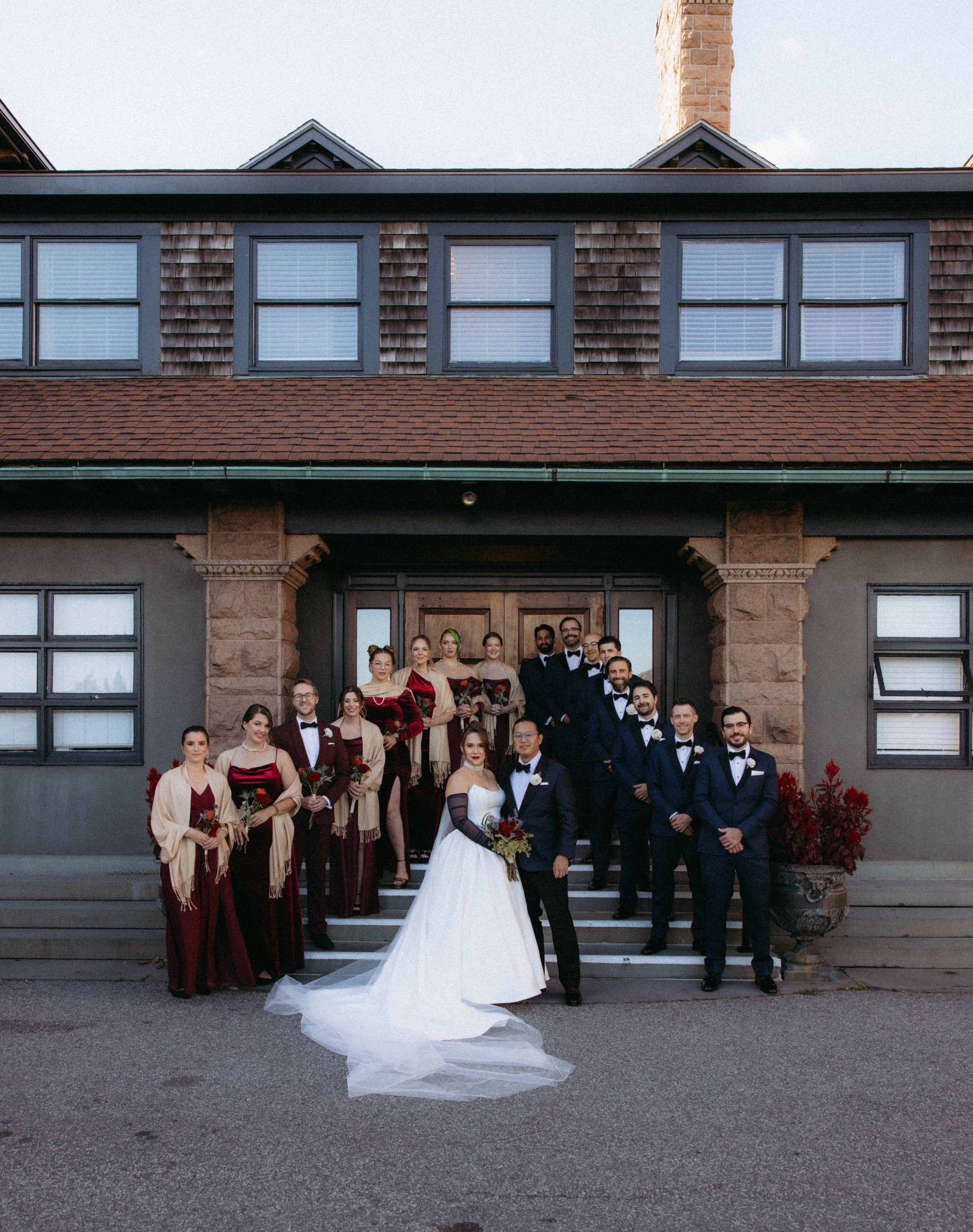 A large group of people dressed in formal attire, including tuxedos and gowns, standing on the steps of a house with a stone and wooden exterior, posing for a wedding photo. The bride is in a white dress with a long train, holding a bouquet, and stan