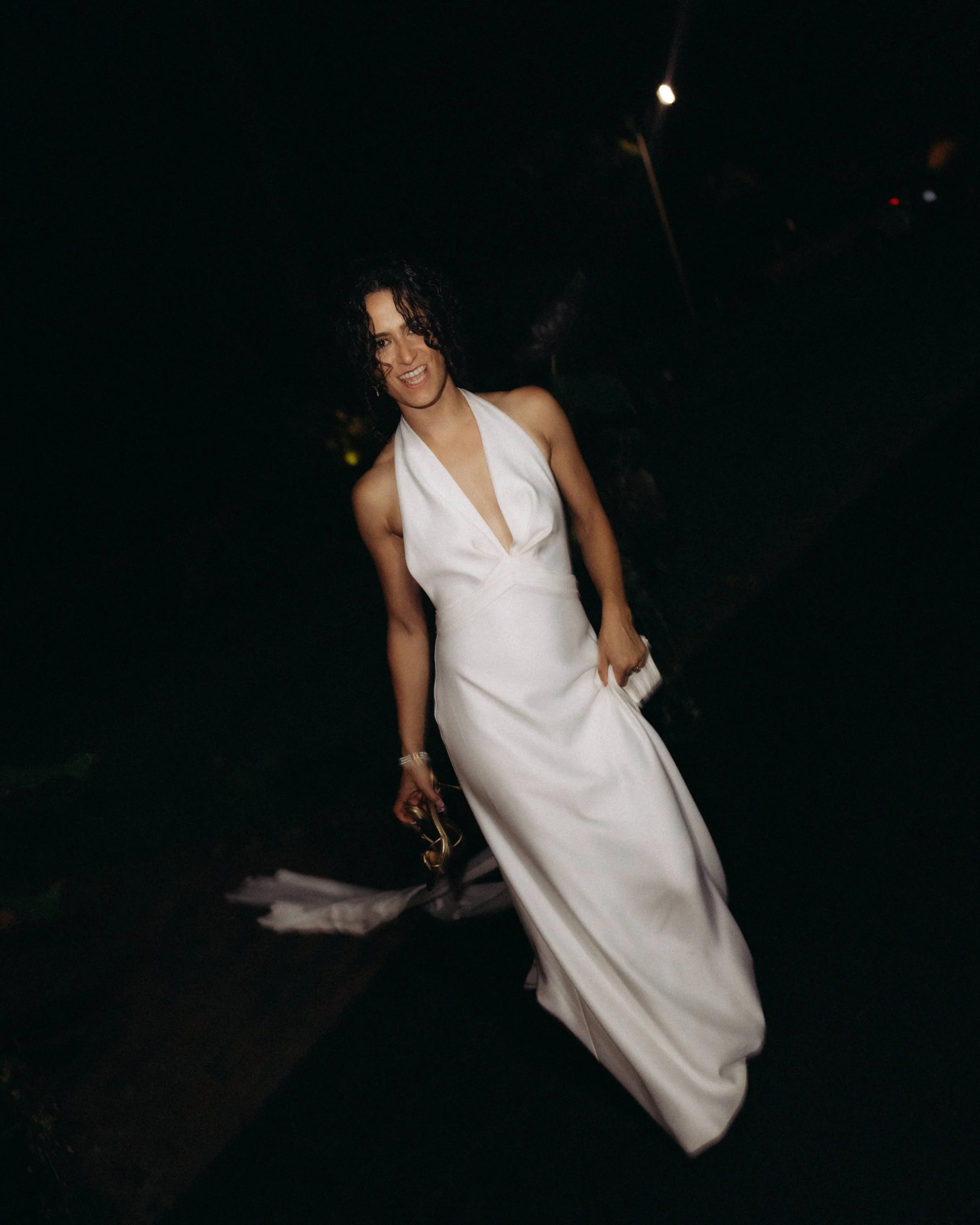 Woman in a white dress standing outdoors at night, smiling and holding sunglasses.