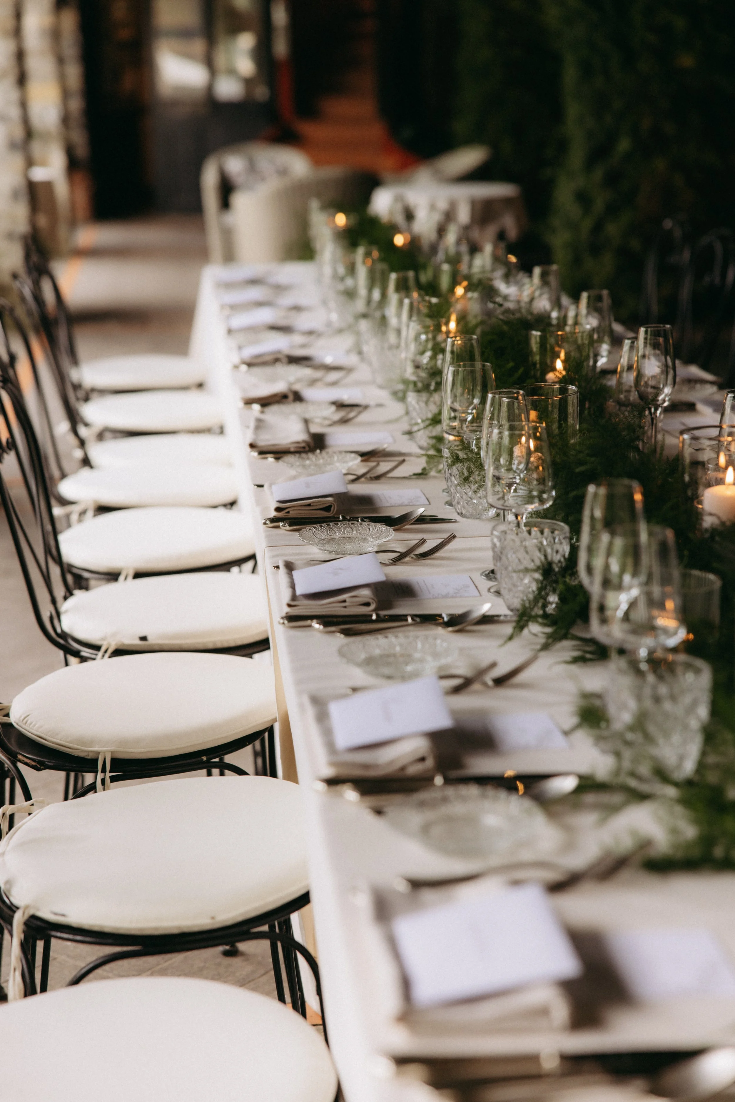 A long dining table set for an elegant meal with white tablecloths, glassware, silverware, and napkins, decorated with greenery and candles, in an outdoor or semi-outdoor setting.