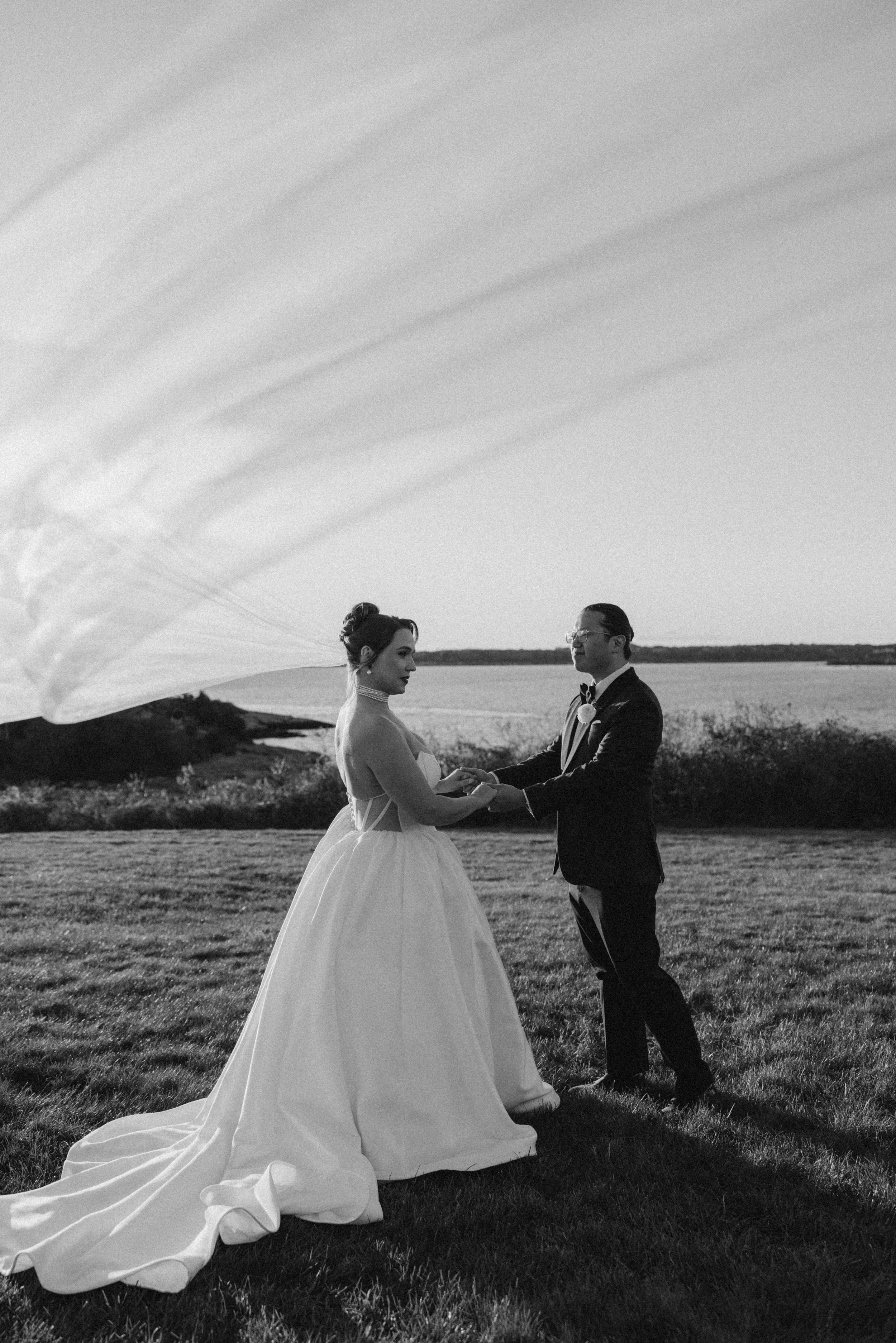 A black and white photo of a bride and groom holding hands outdoors by a body of water, with a large leaf partially covering the sky.
