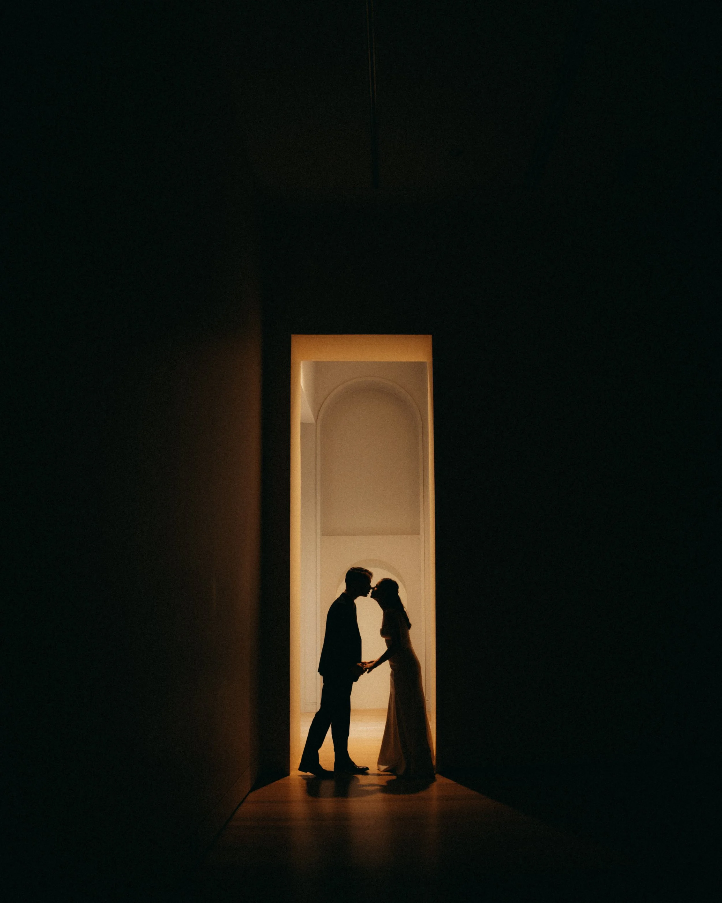 A silhouette of a couple holding hands, standing close and facing each other, inside a dark room with a tall, illuminated rectangular archway behind them. The background is black, highlighting the figures and the bright archway.