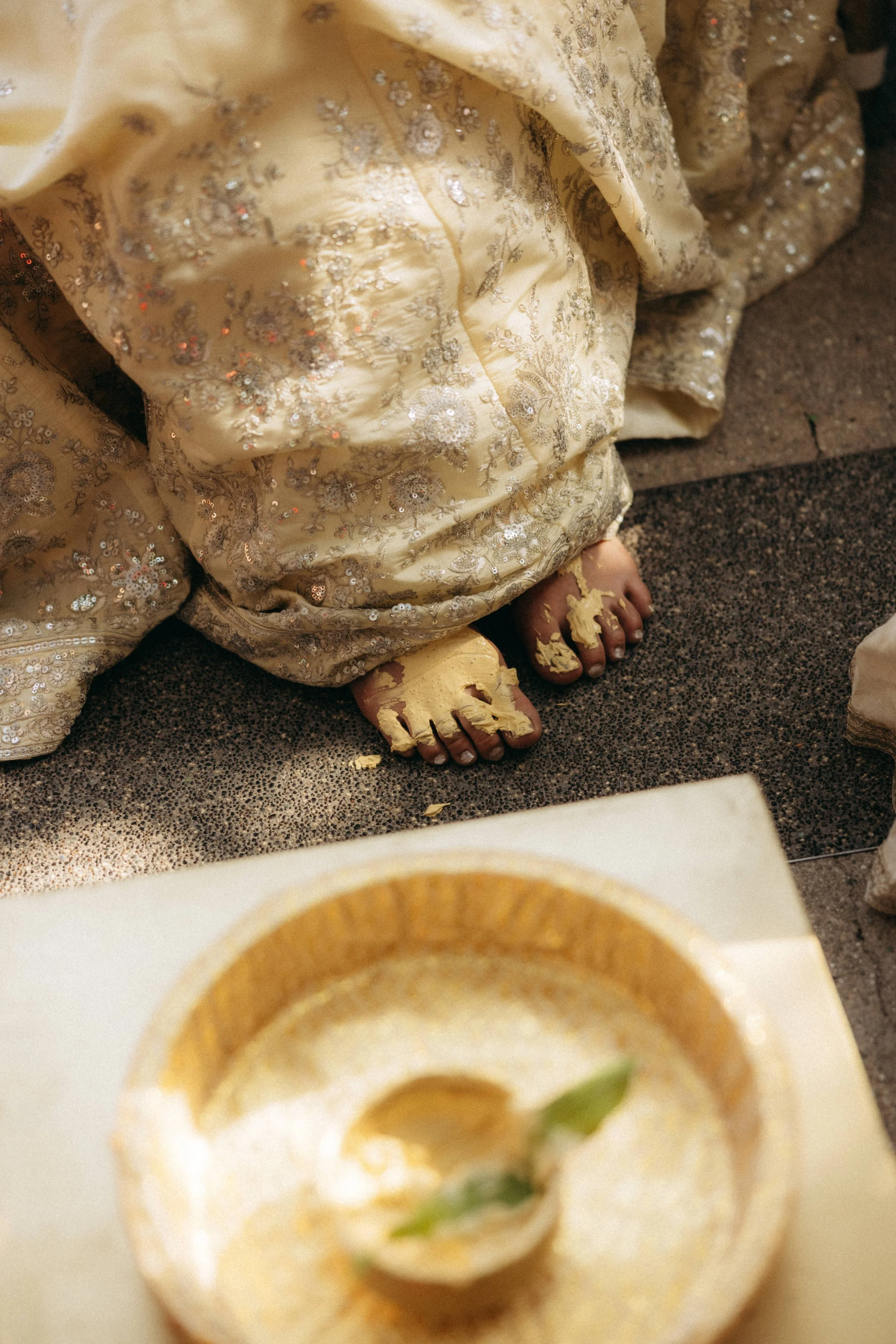 Close-up of a person in embroidered clothing with gold and silver sequins and beads, sitting on a carpeted floor, with shiny gold sandals on their feet, with some peeled paint on their toes.