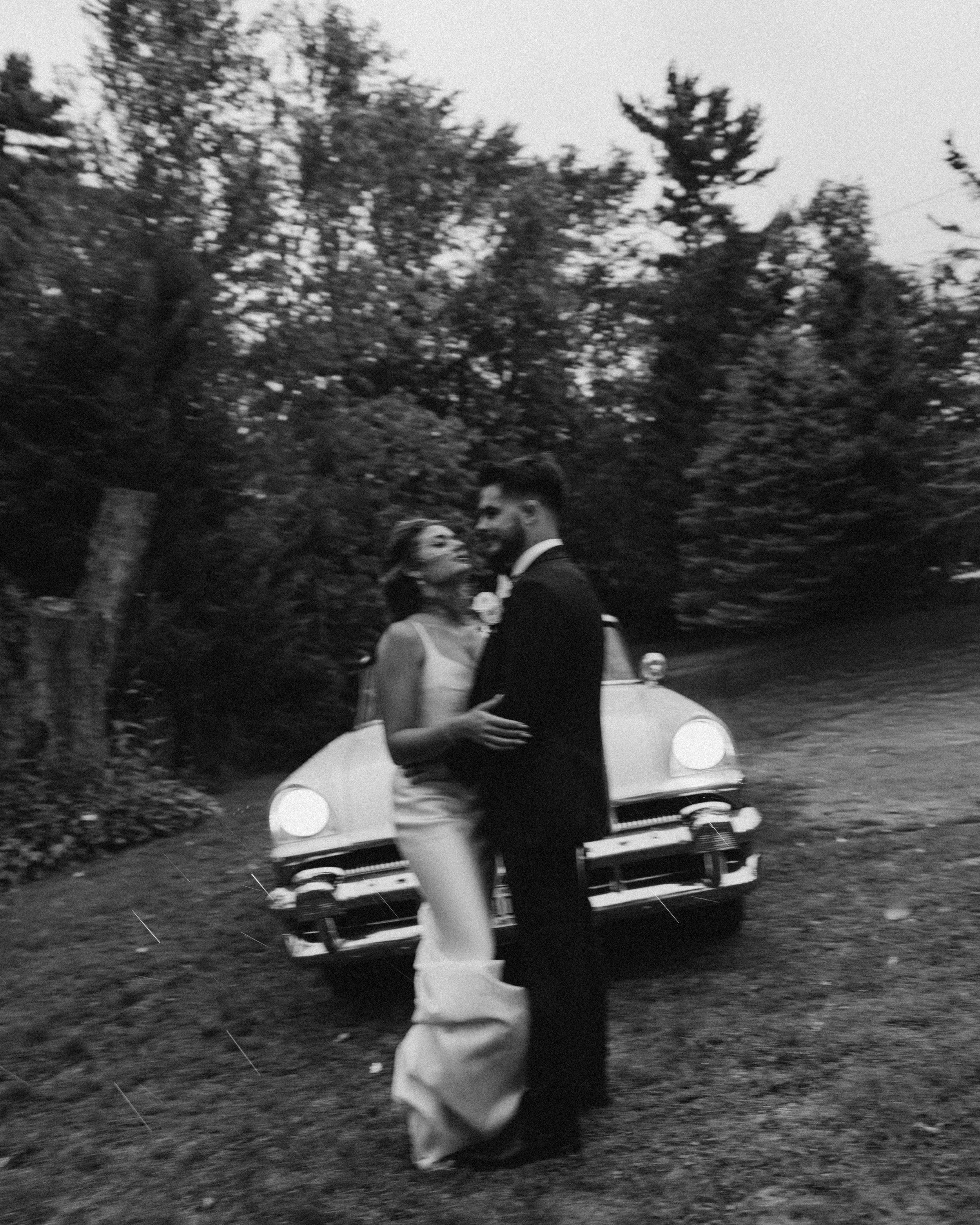 A black and white photo of a couple in wedding attire, dancing outdoors at sunset or dusk. The woman wears a white wedding gown with a veil, and the man wears a dark suit. They are looking into each other's eyes, with trees and a vintage white car in