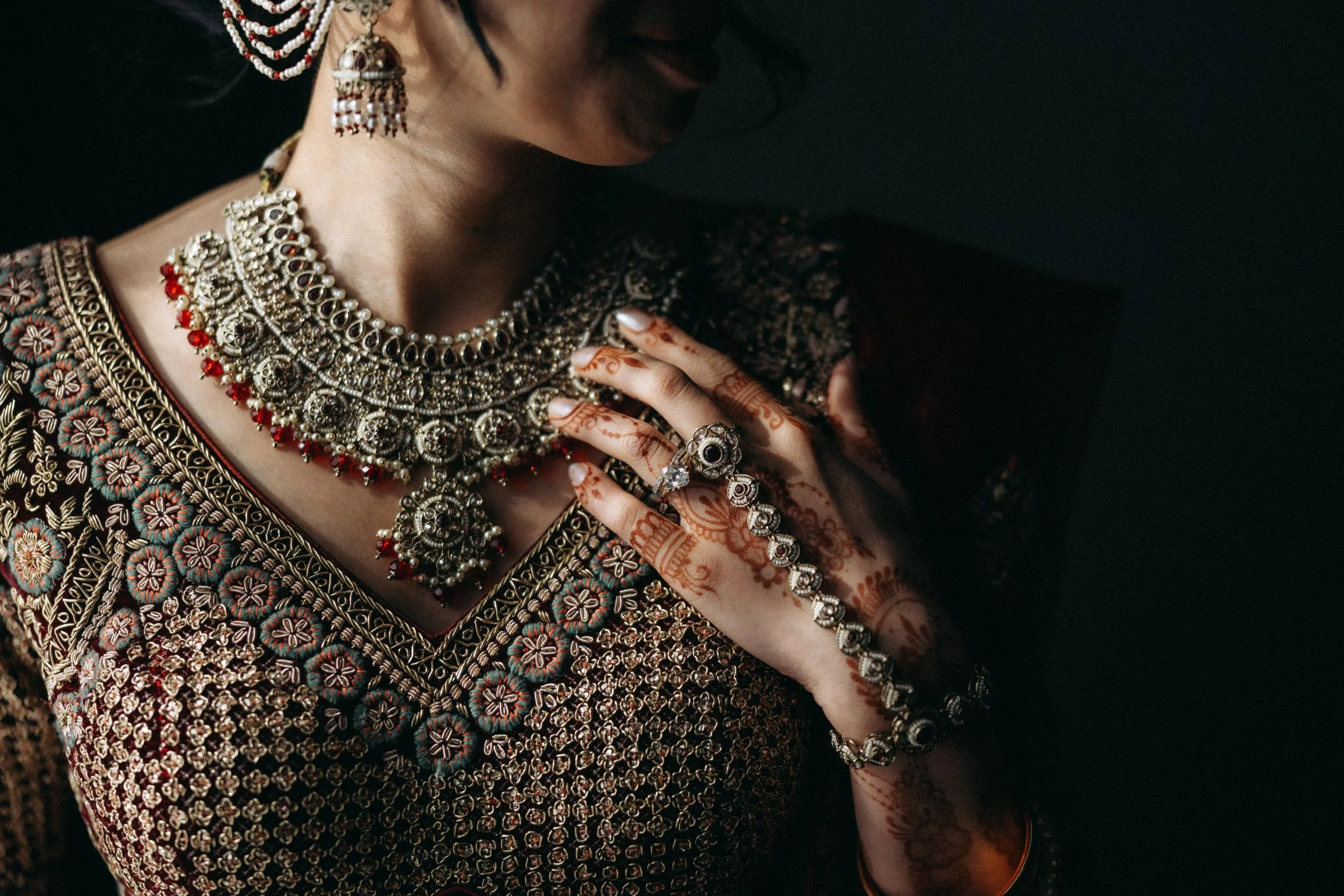 Close-up of a woman wearing traditional Indian jewelry, including a large silver necklace with red beads, matching earrings, rings, and a bracelet. She has henna designs on her hands and is dressed in a heavily embroidered dark-colored outfit with fl