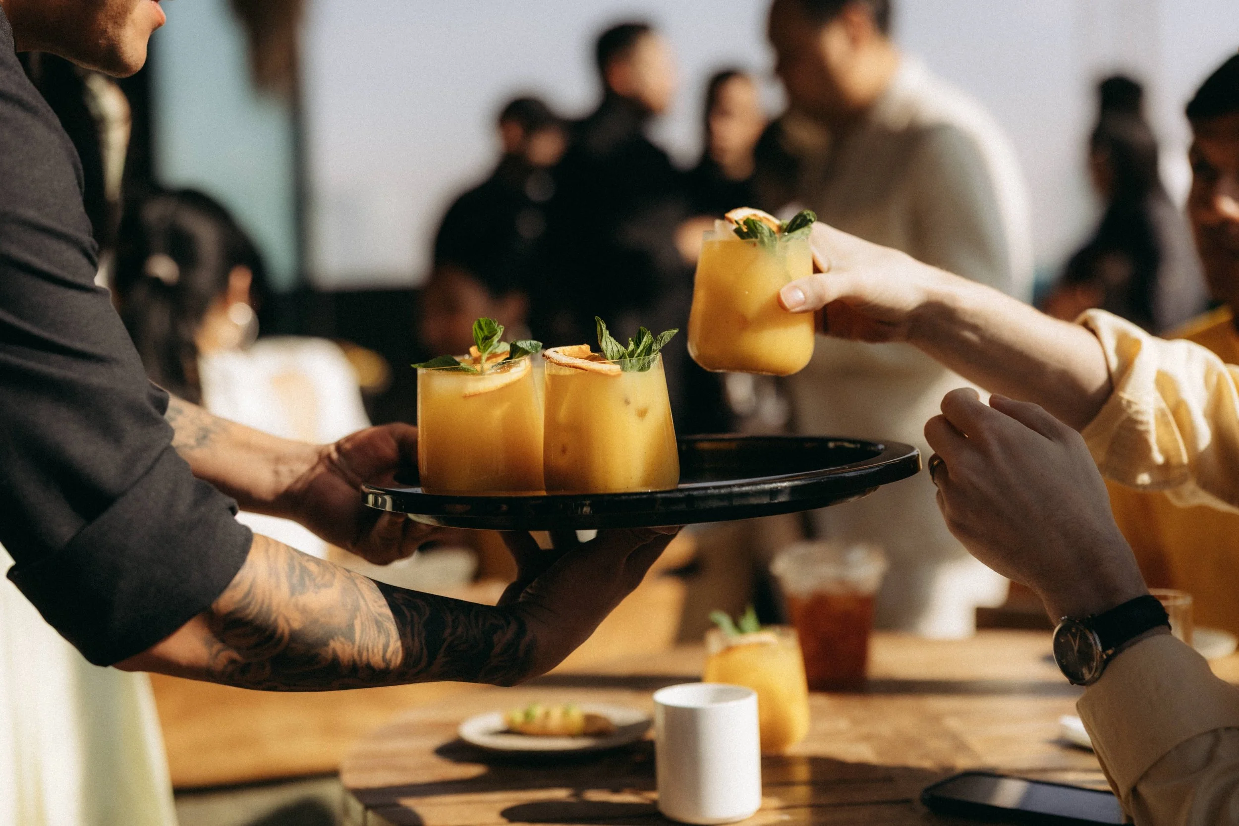 A person with tattooed arms is holding a black tray with three glasses of yellow-orange cocktail garnished with green herbs, and another person is handing a similar glass to them in a social setting with blurred people in the background.