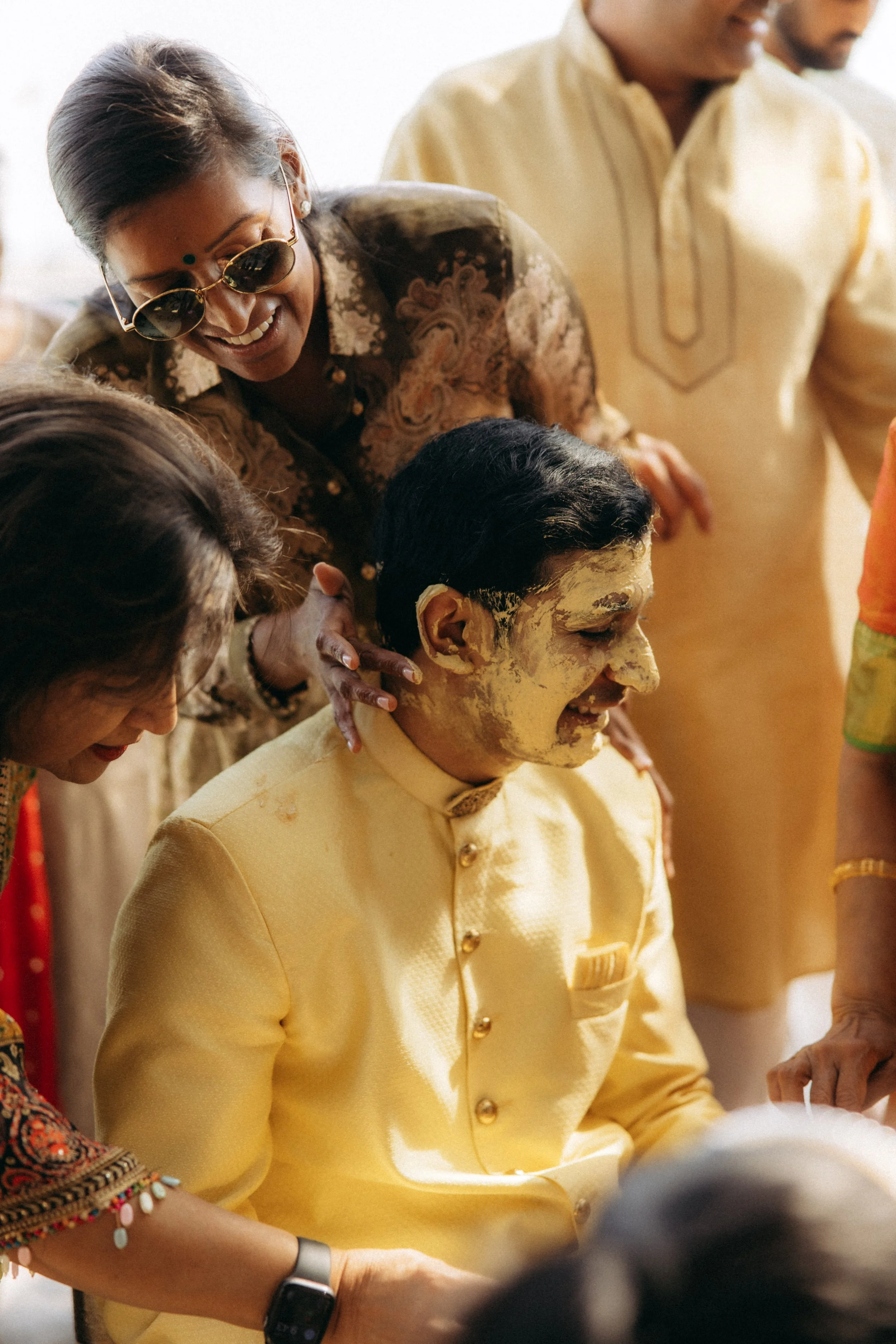 A woman with sunglasses and a traditional patterned shirt is smiling and assisting a man in a yellow traditional outfit with a yellow paste on his face. Other people are partially visible in the background.