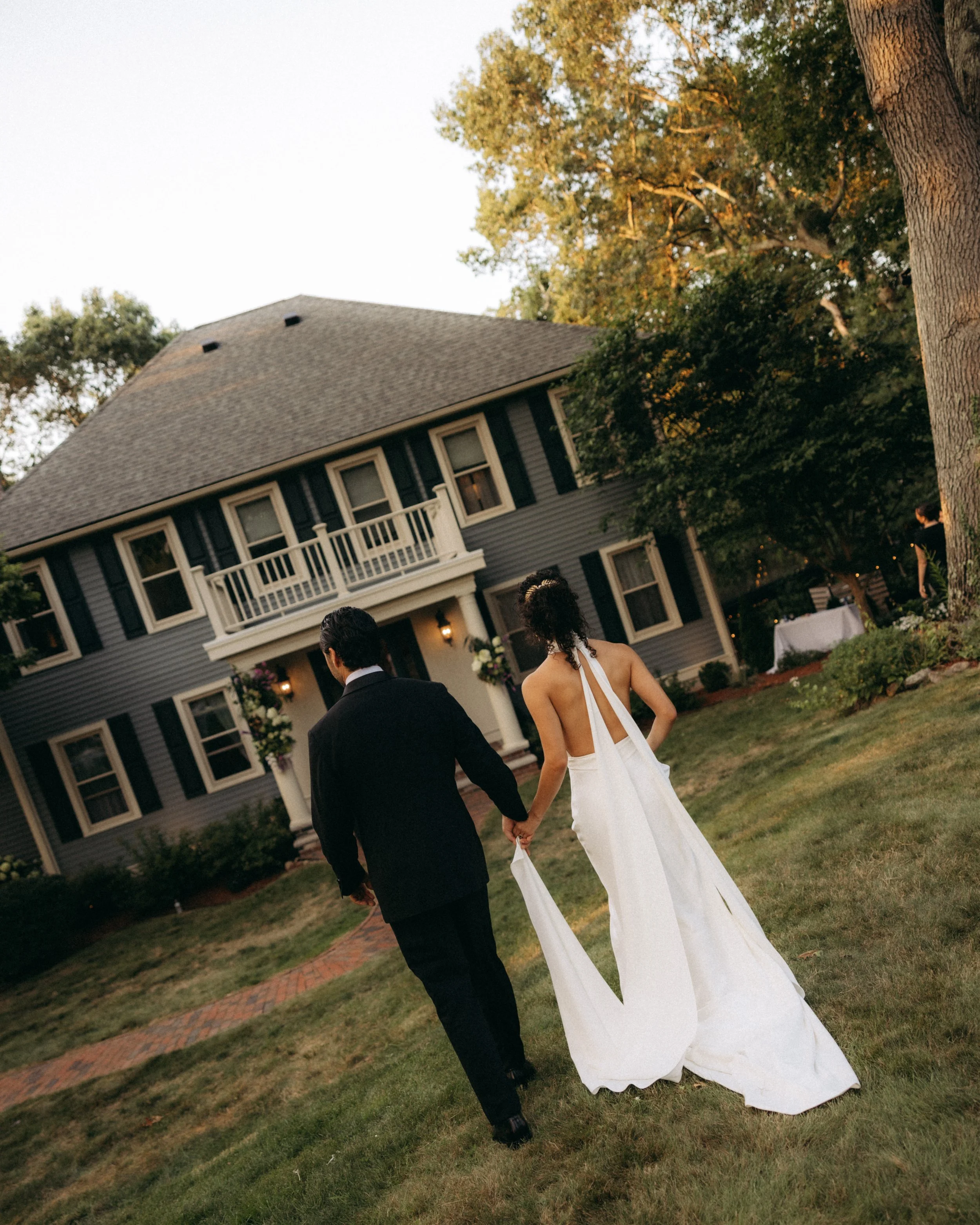 A bride and groom walk hand in hand in front of a large house with a porch and black shutters, surrounded by trees with green and golden leaves, during sunset.