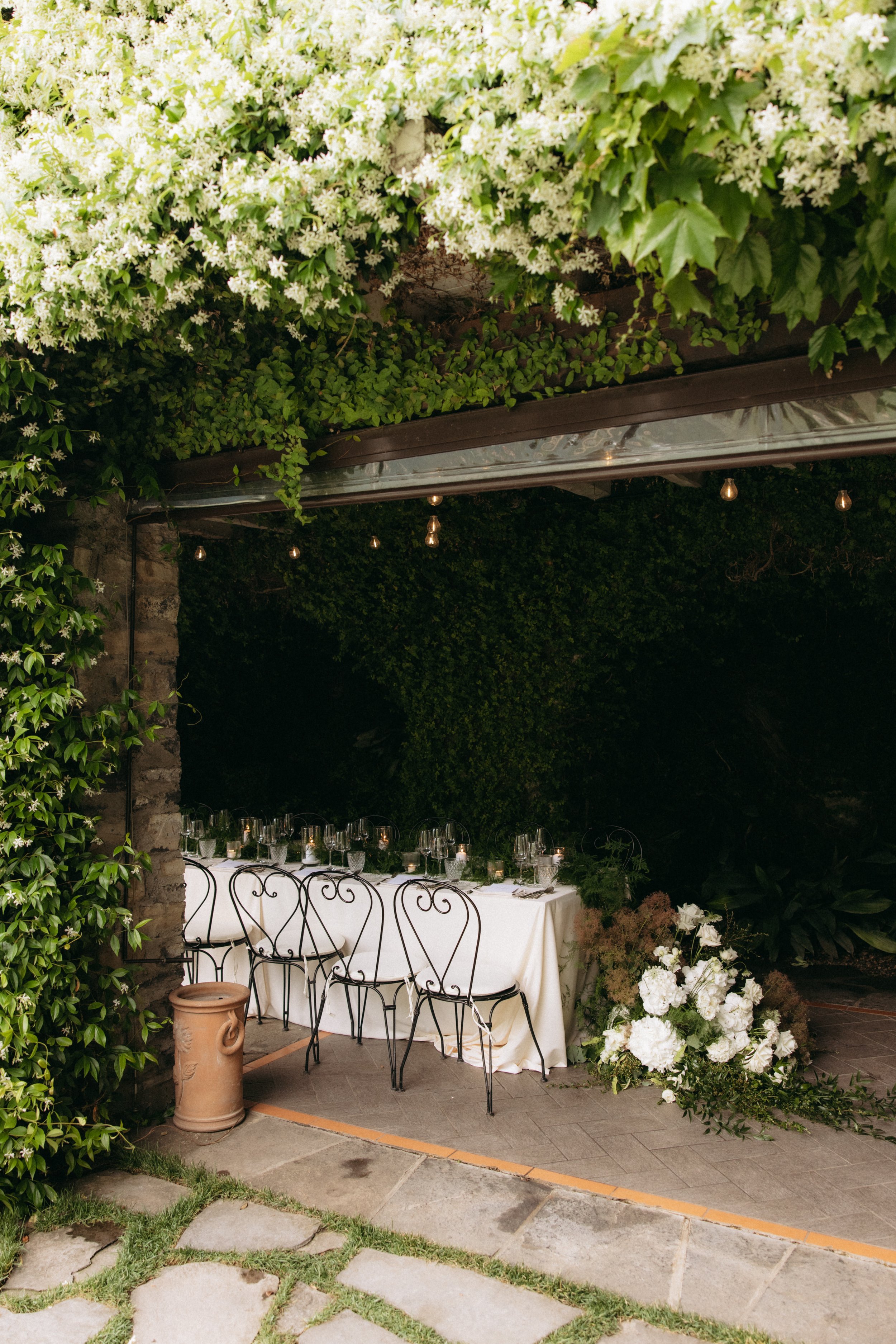 A long, elegant outdoor dining table set with white tablecloth, glassware, and candles, surrounded by vintage-style black chairs, under a canopy of green leaves and hanging string lights, creating a festive atmosphere for a celebration.
