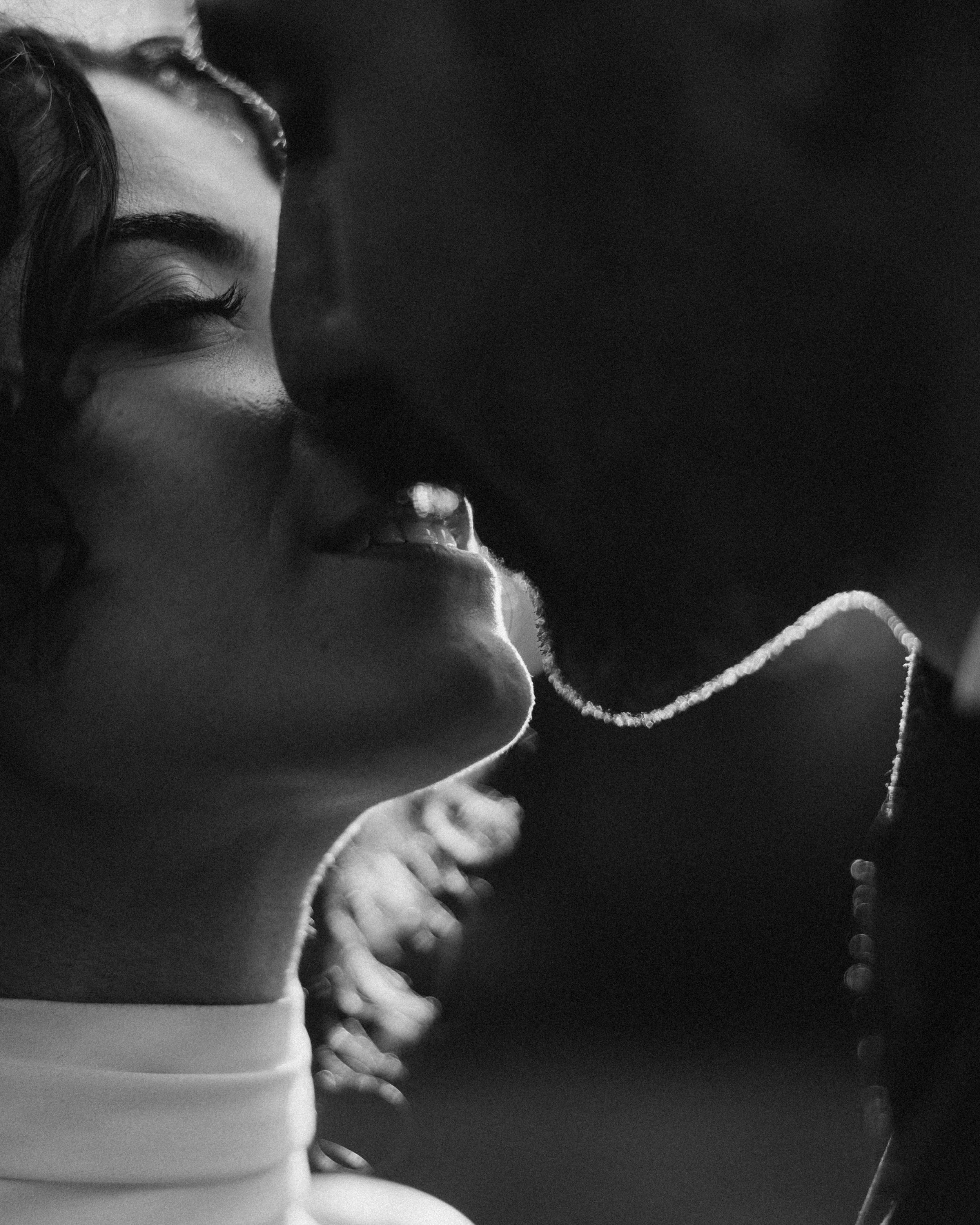 A black and white close-up photo of two people about to kiss, capturing the side profiles of their faces, along with jewelry and jewelry chains.