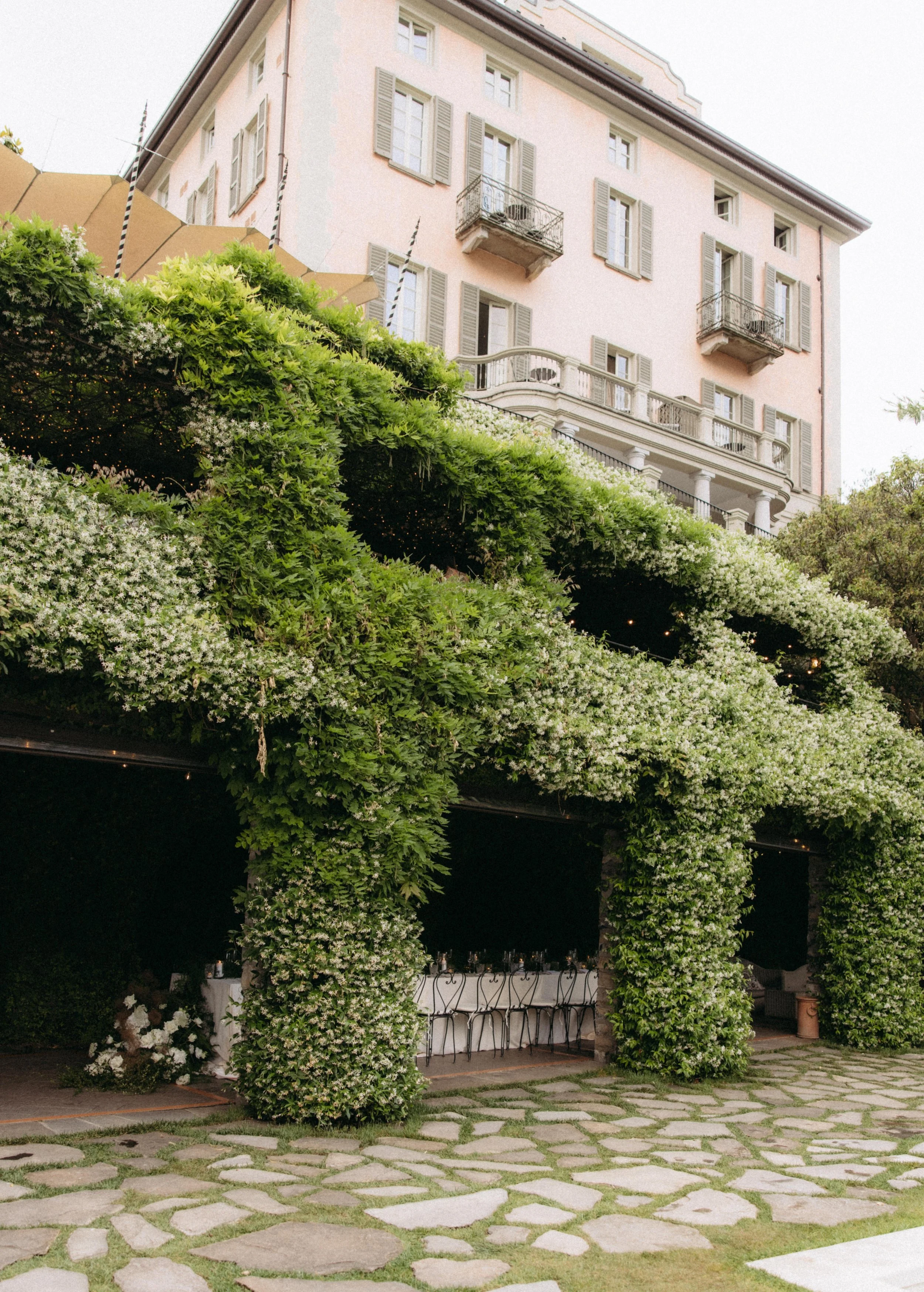 Exterior view of a pink multi-story house with gray shutters and small balconies, partially obscured by a large green arbor decorated with string lights, under an overcast sky.