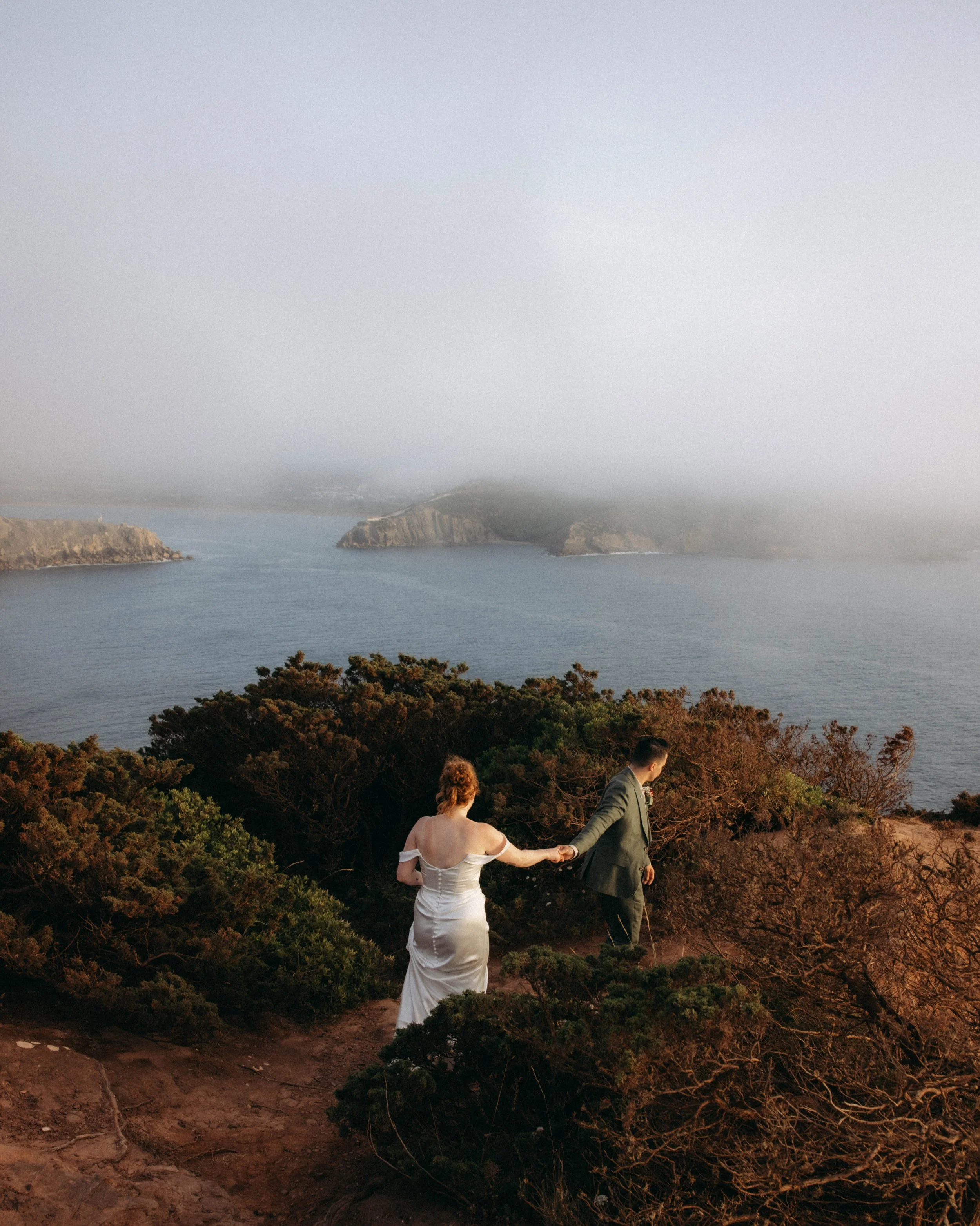 A bride and groom holding hands and walking along a trail on a hillside with a view of the water and distant cliffs and foggy sky in the background.
