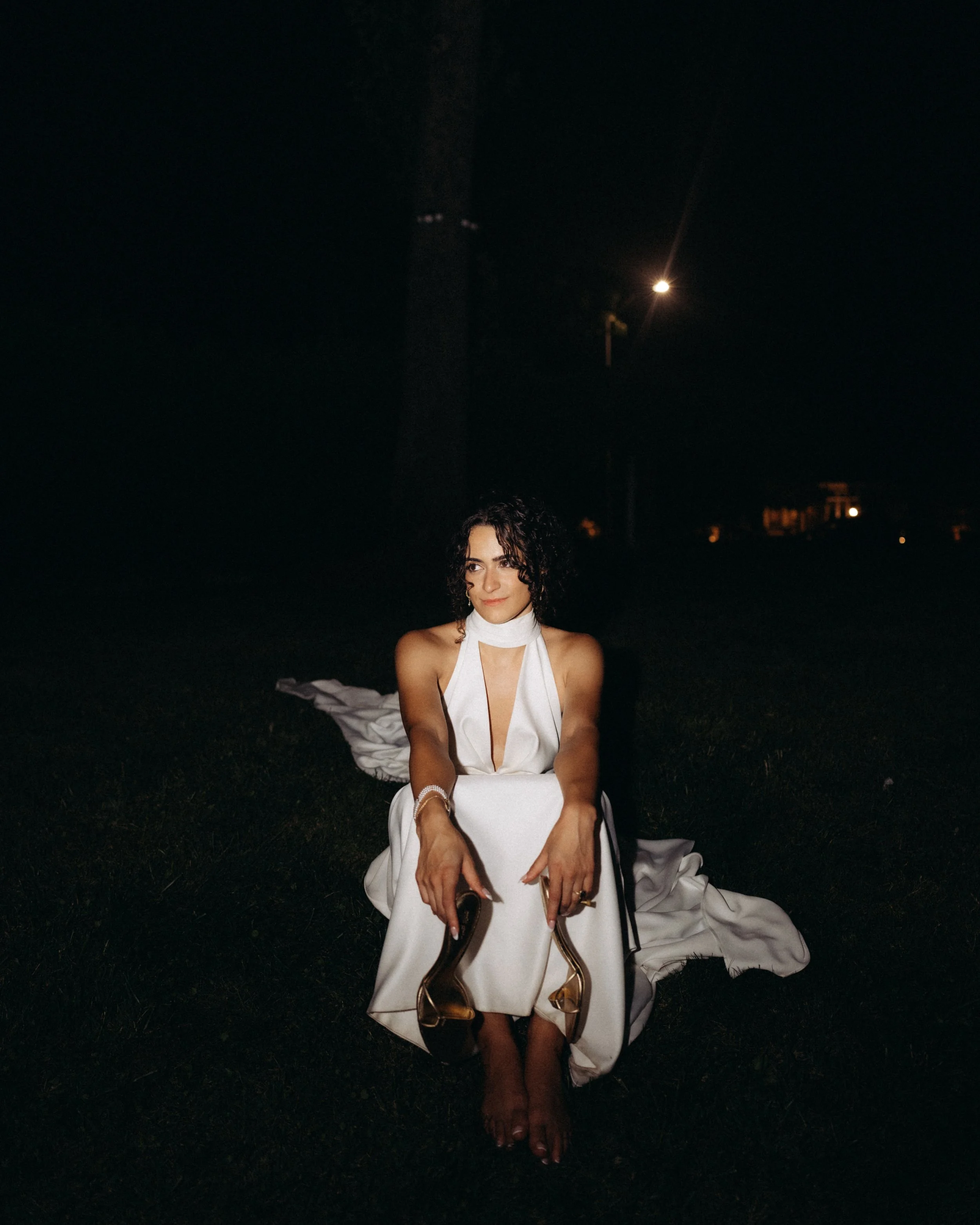 A woman with curly dark hair and light skin is sitting outdoors at night. She is wearing a white dress with a deep neckline, jewelry, and heels. She is holding sunglasses in her hands, and her feet are bare on the grass.