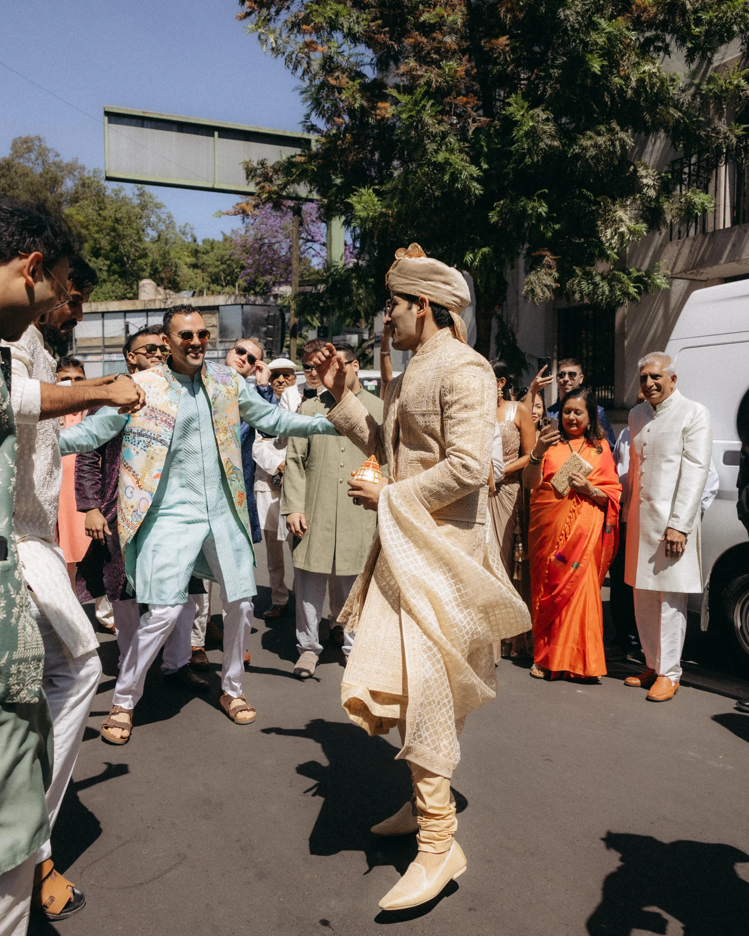 A group of people dressed in traditional Indian attire participating in a cultural celebration outdoors under a blue sky, with trees and a building in the background. A man in a cream-colored sherwani and turban appears to be performing or leading a 