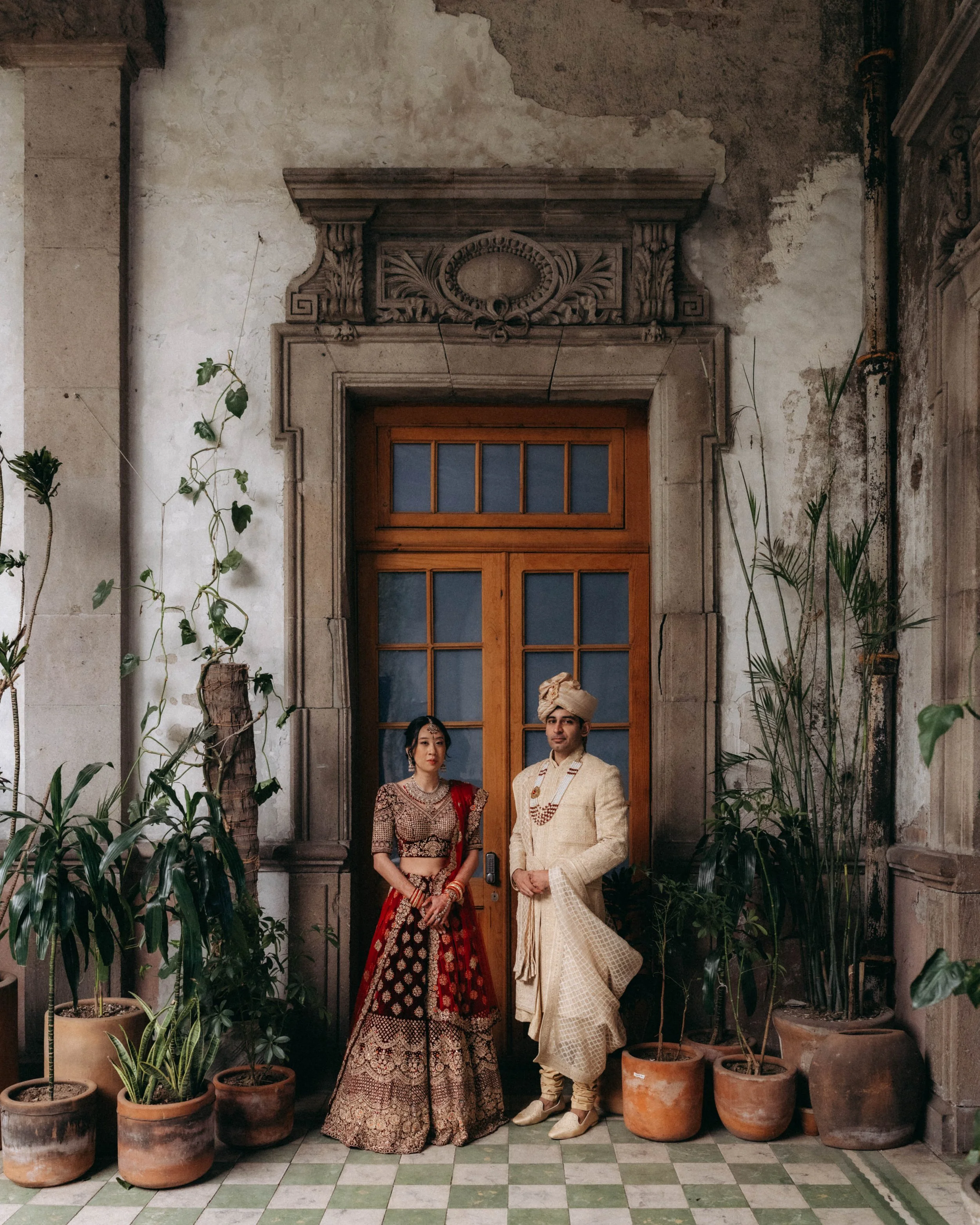 A man and a woman dressed in traditional South Asian attire standing in front of a rustic wooden door with faded decorative architecture above it. The woman is wearing a maroon and gold lehenga choli with jewelry, and the man is dressed in a cream-co