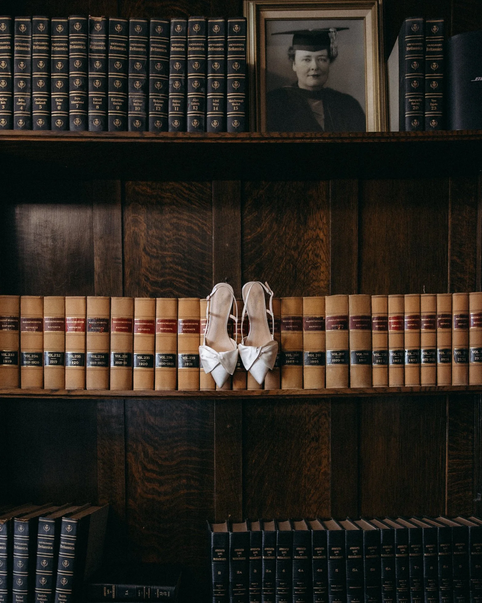 A wooden bookshelf with several volumes of Britannica Encyclopedia on the top and bottom shelves, a framed black and white photograph of a woman in a graduation cap and gown on the higher shelf, and a pair of beige high-heeled shoes with a bow placed