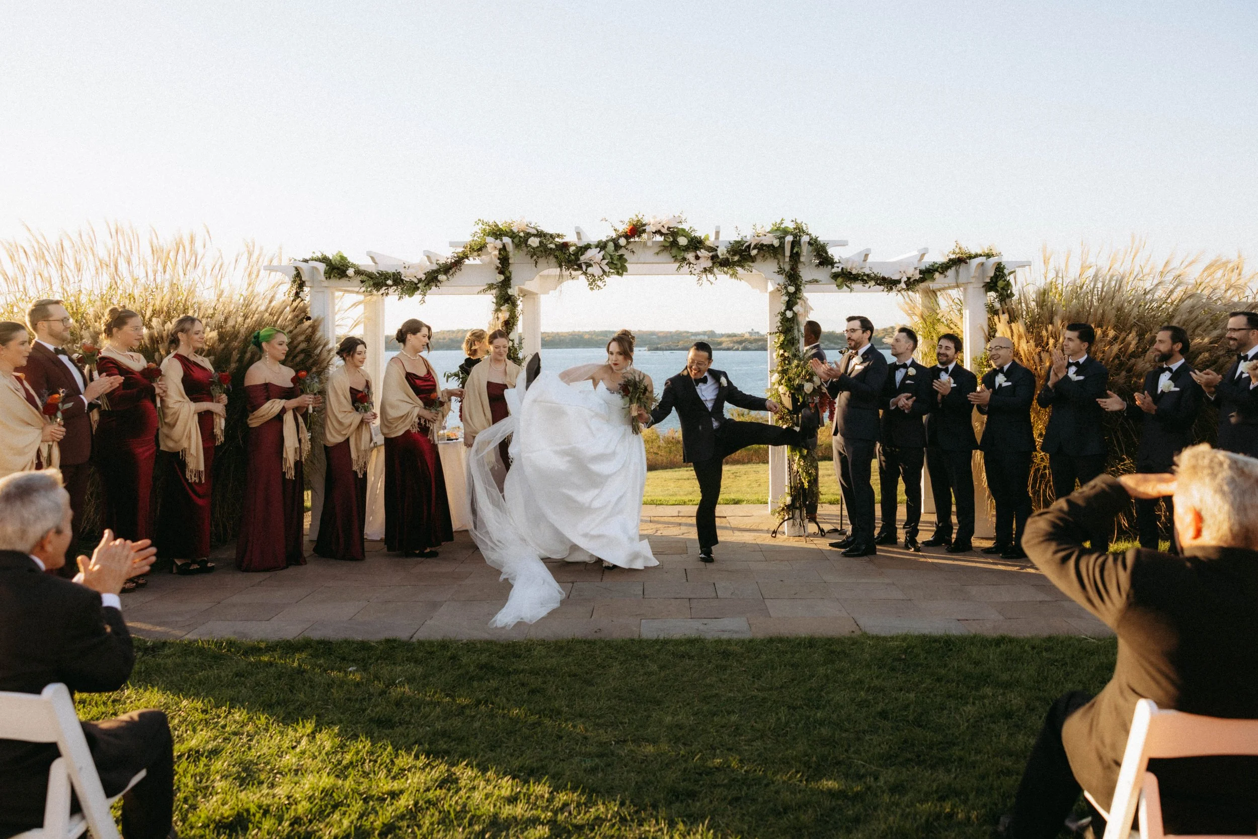 A wedding ceremony outdoors with a couple kissing under a decorated archway, surrounded by friends and family clapping and celebrating.