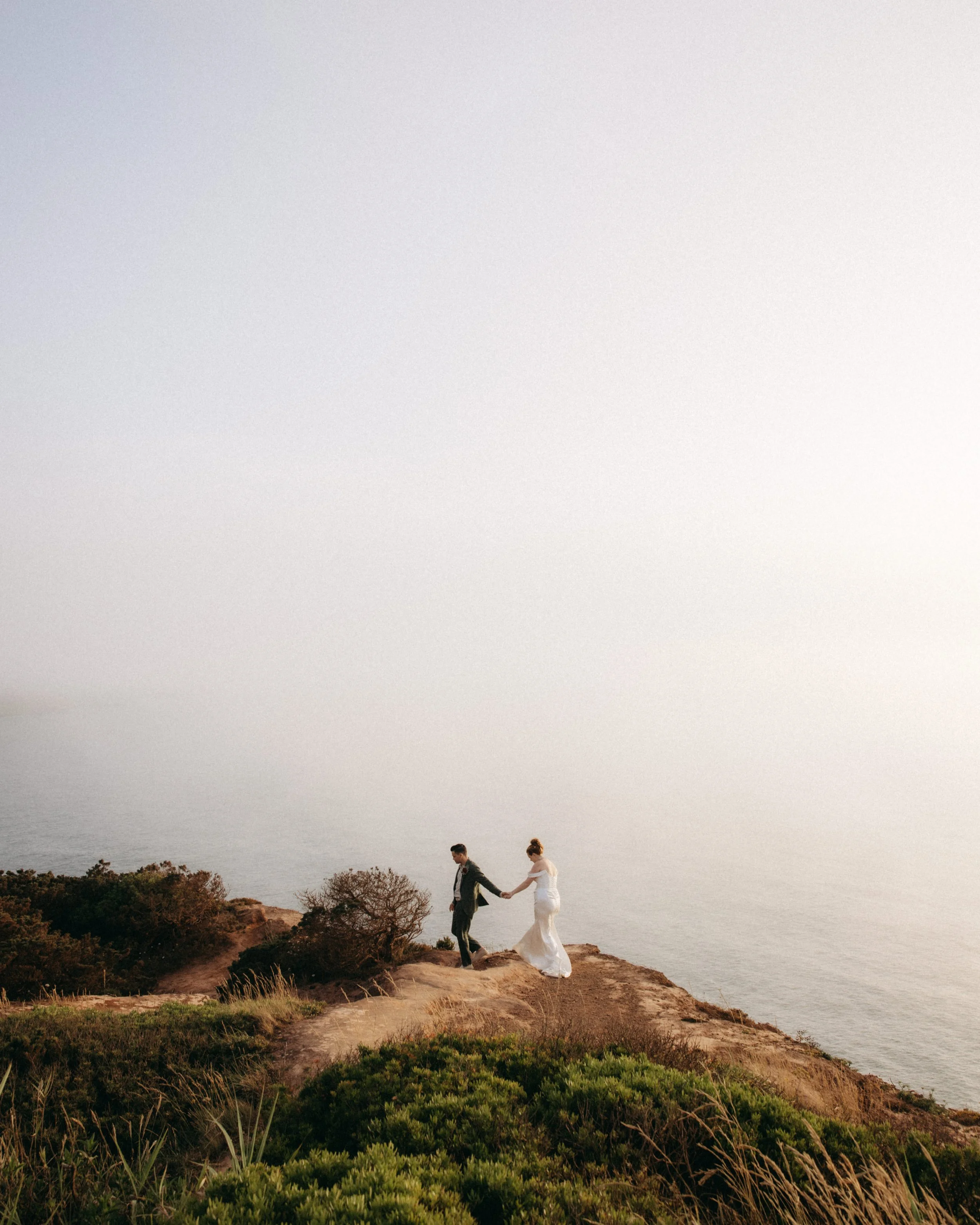 A couple walking hand-in-hand along a cliff edge overlooking the ocean during sunset.