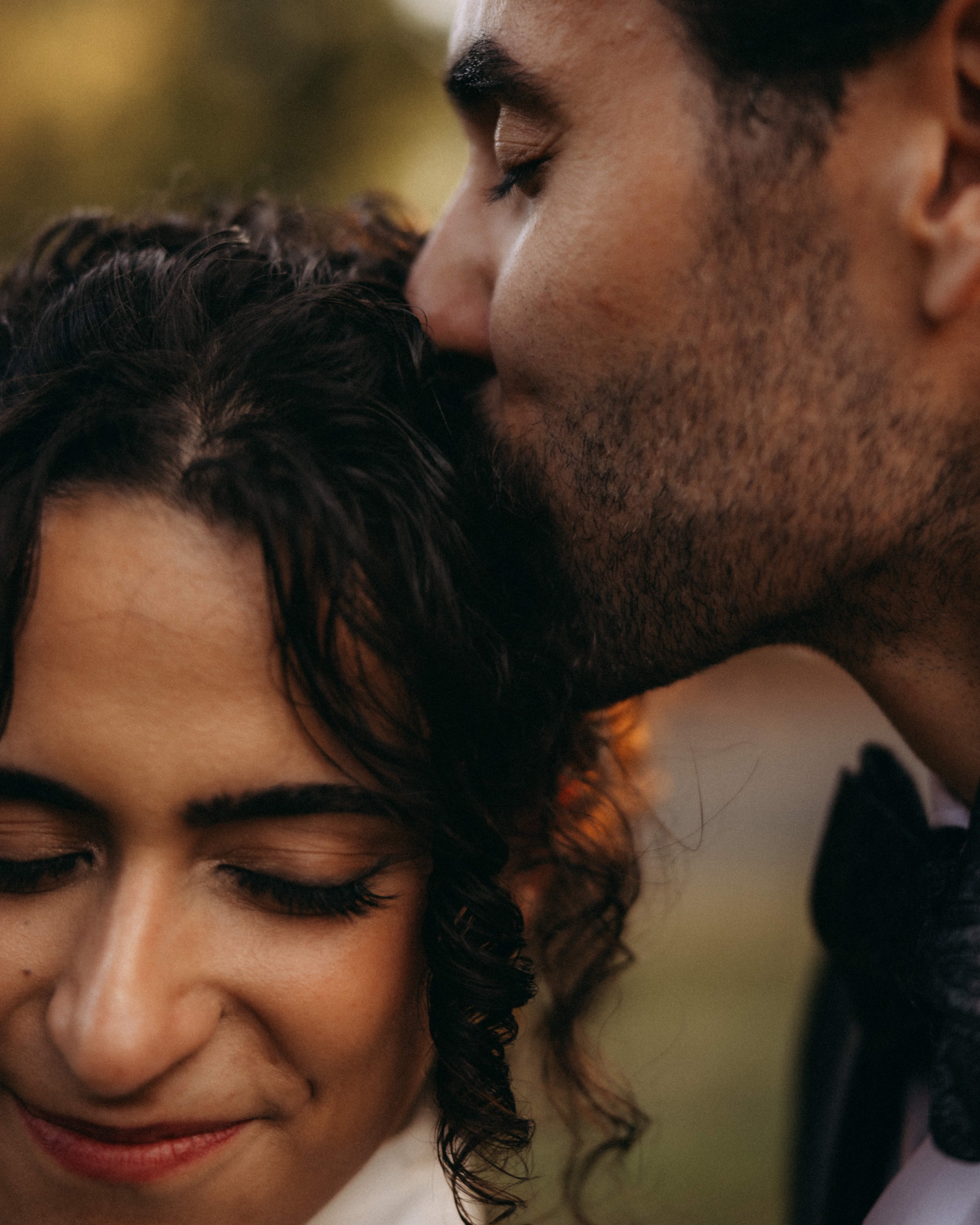 A woman with dark, curly hair gently nuzzling a man's cheek. The woman has a warm smile and her eyes are closed. The man has a beard and is leaning his face into the woman's touch, with his eyes also closed. The background is blurred and warm-toned.