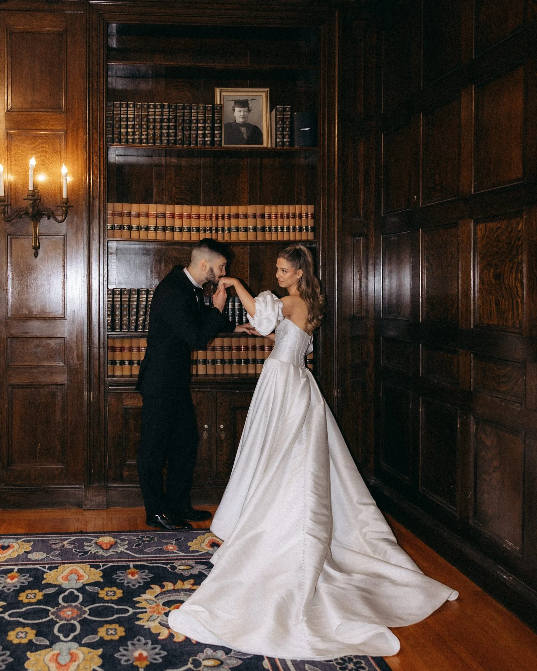 A bride and groom are holding hands and looking at each other in a wooden-paneled room with a bookshelf, a framed photograph of a woman in academic attire, and a floral rug.