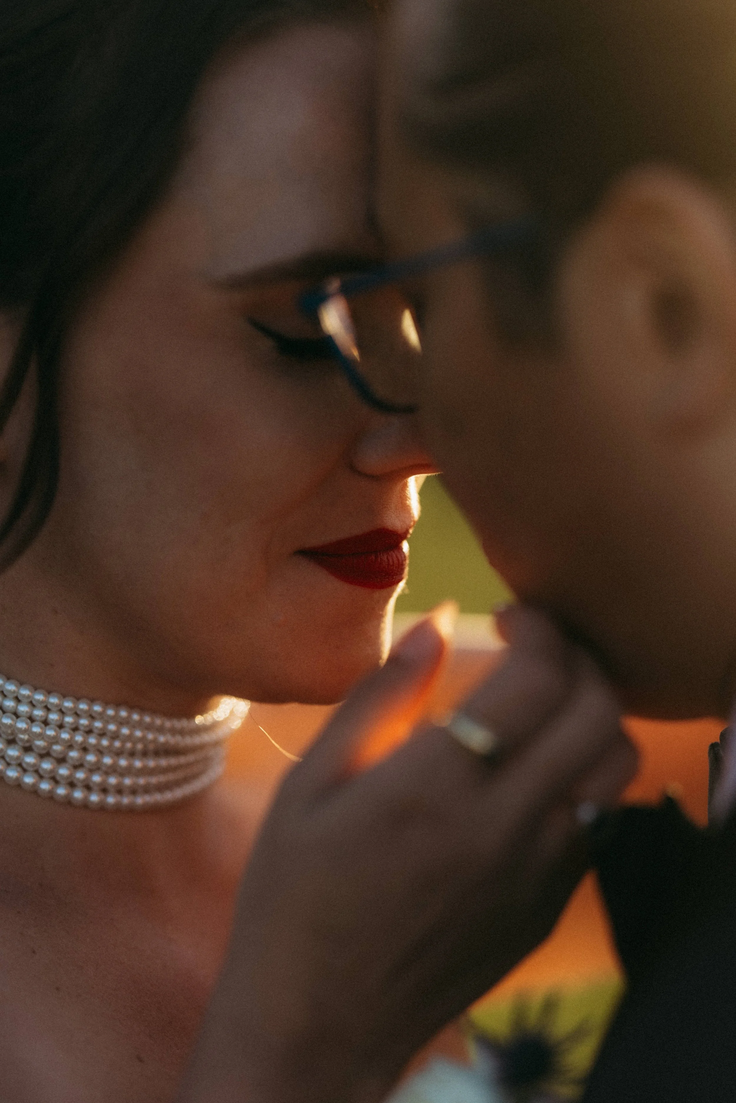 Close-up of a woman with dark hair wearing glasses, red lipstick, and a pearl necklace, looking at her phone.