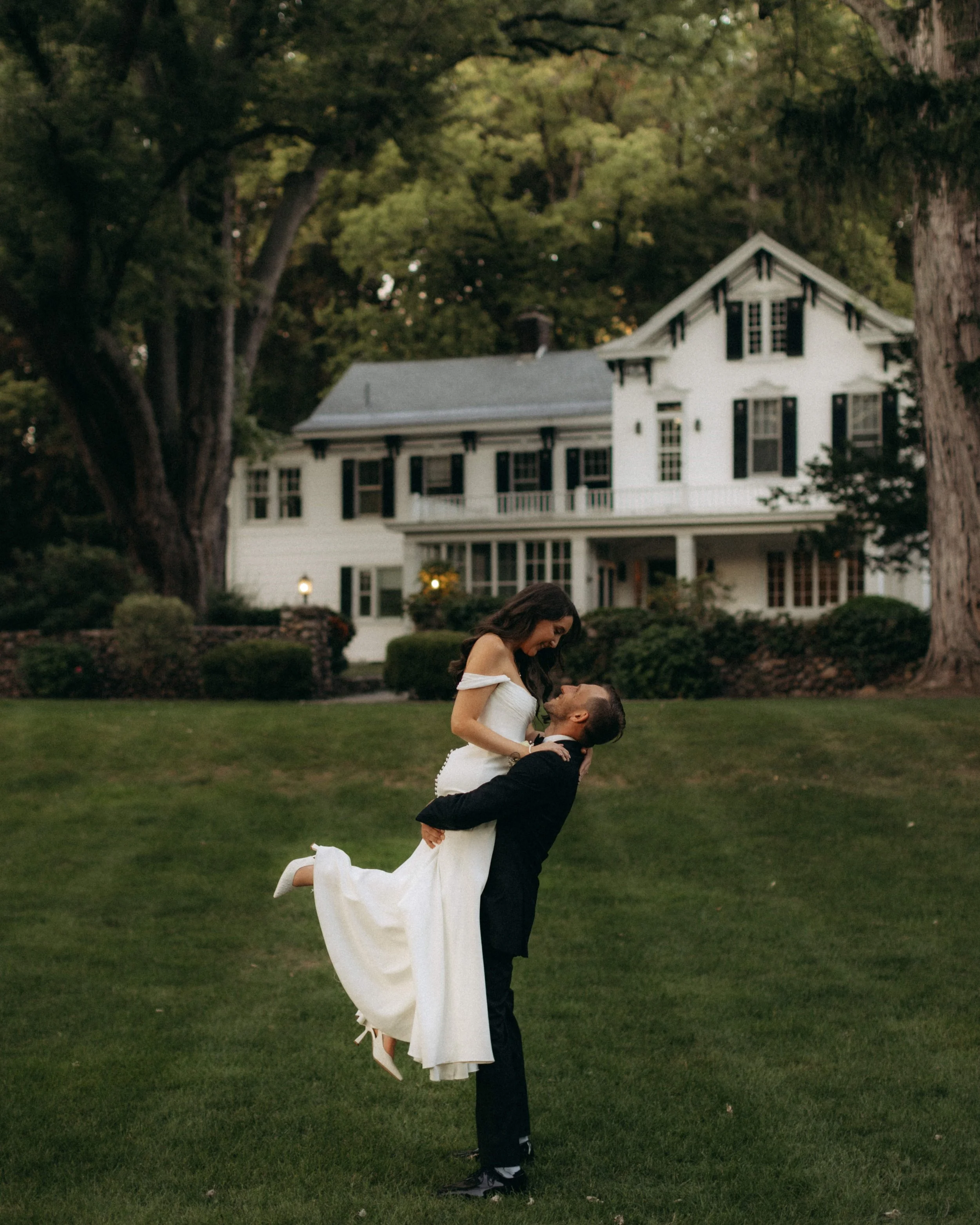 A man holding a woman in the air on a lawn in front of a large white house with black shutters, surrounded by trees.