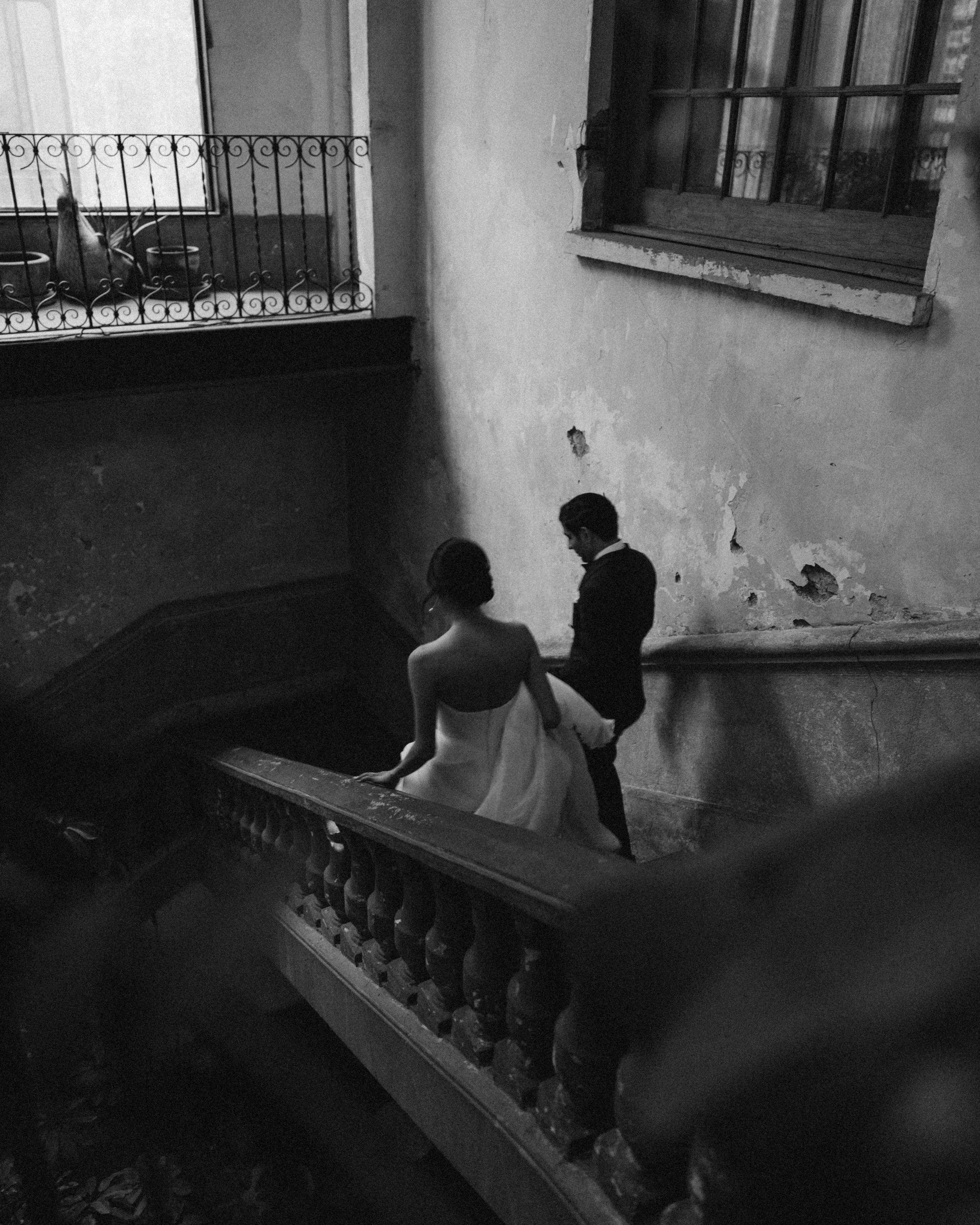 Black and white photo of three people sitting on a staircase in a building with peeling paint wall and a window. Two men in dark suits are talking to a woman in a light-colored dress. The staircase has a metal railing with curved patterns.